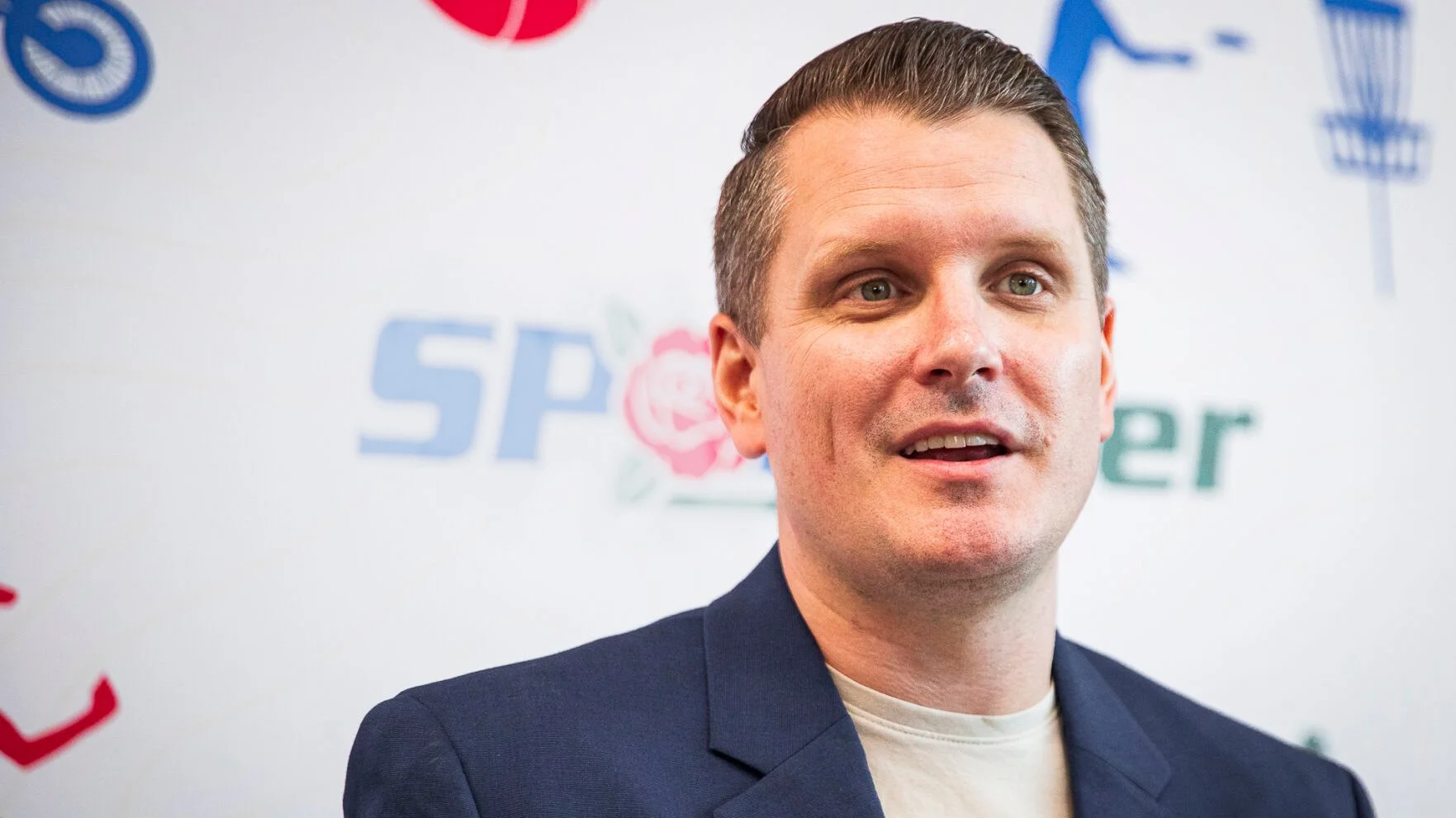 Greg Tepper speaking at a sports media event, wearing a navy blazer and white shirt, standing in front of a branded backdrop.