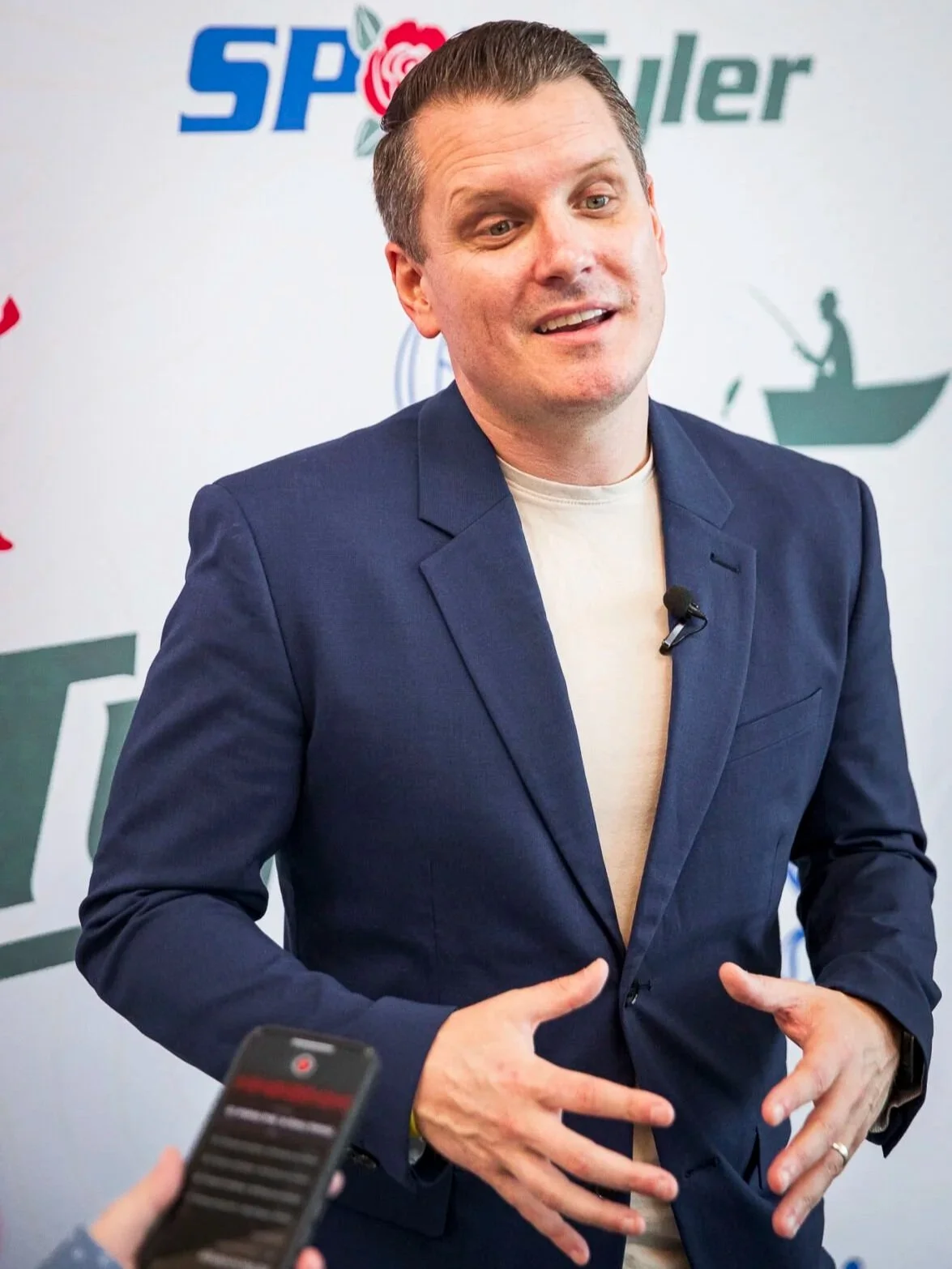 Greg Tepper presenting at a media event, wearing a navy blazer and speaking on stage with a lapel microphone.