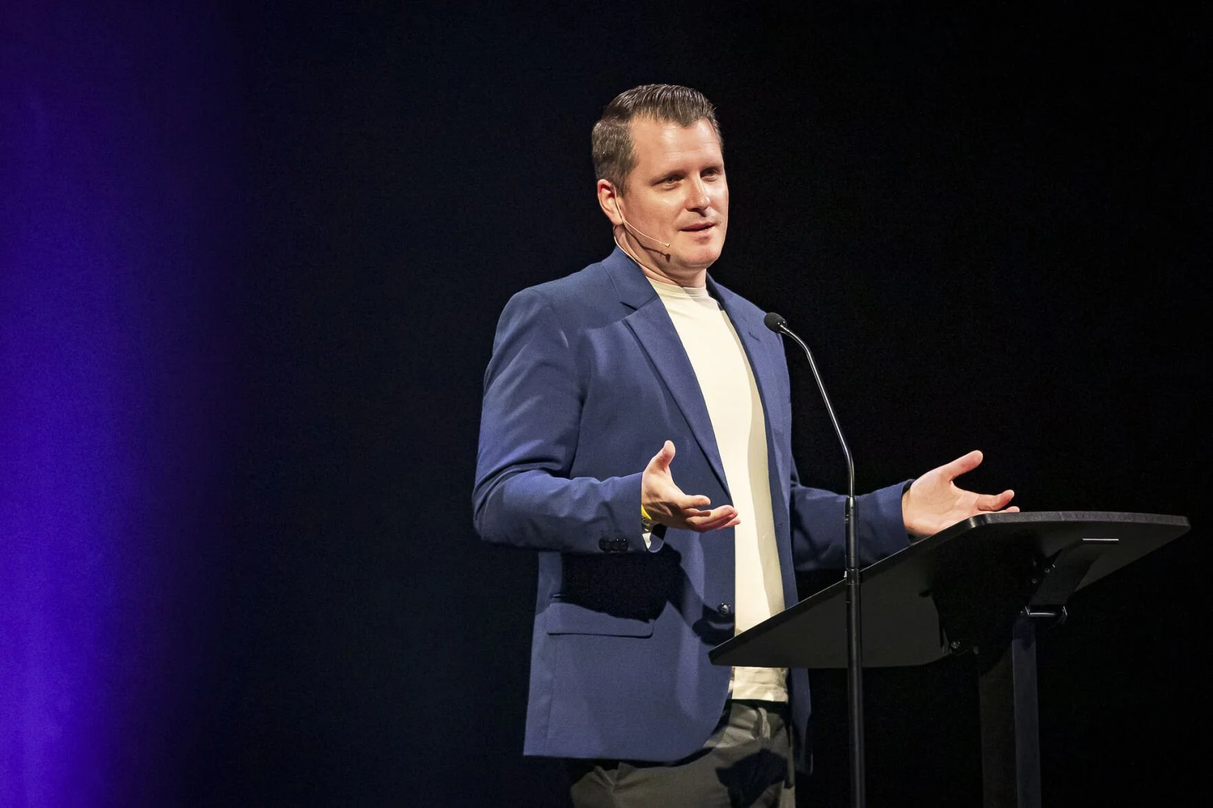 Man giving a presentation on stage behind a lectern, wearing a navy blazer and a cream-colored shirt, with a microphone, gesturing with hands against a dark background.