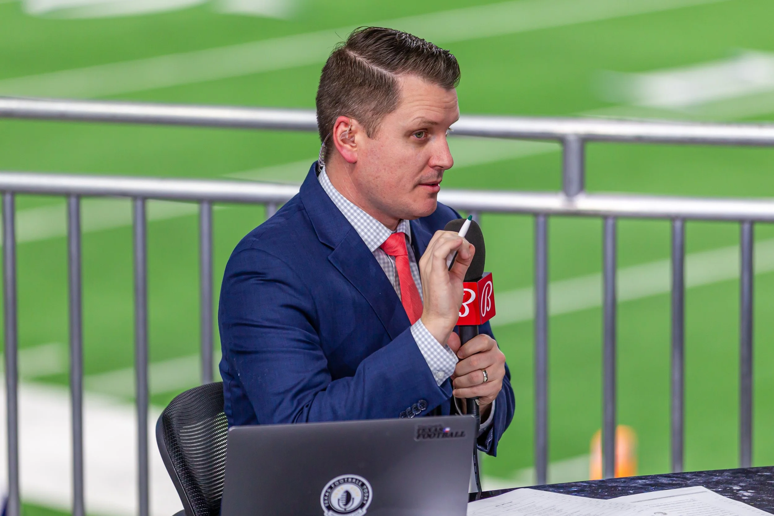 Greg Tepper hosting a live broadcast, wearing a blue suit and pink tie, speaking into a microphone at a sporting event.