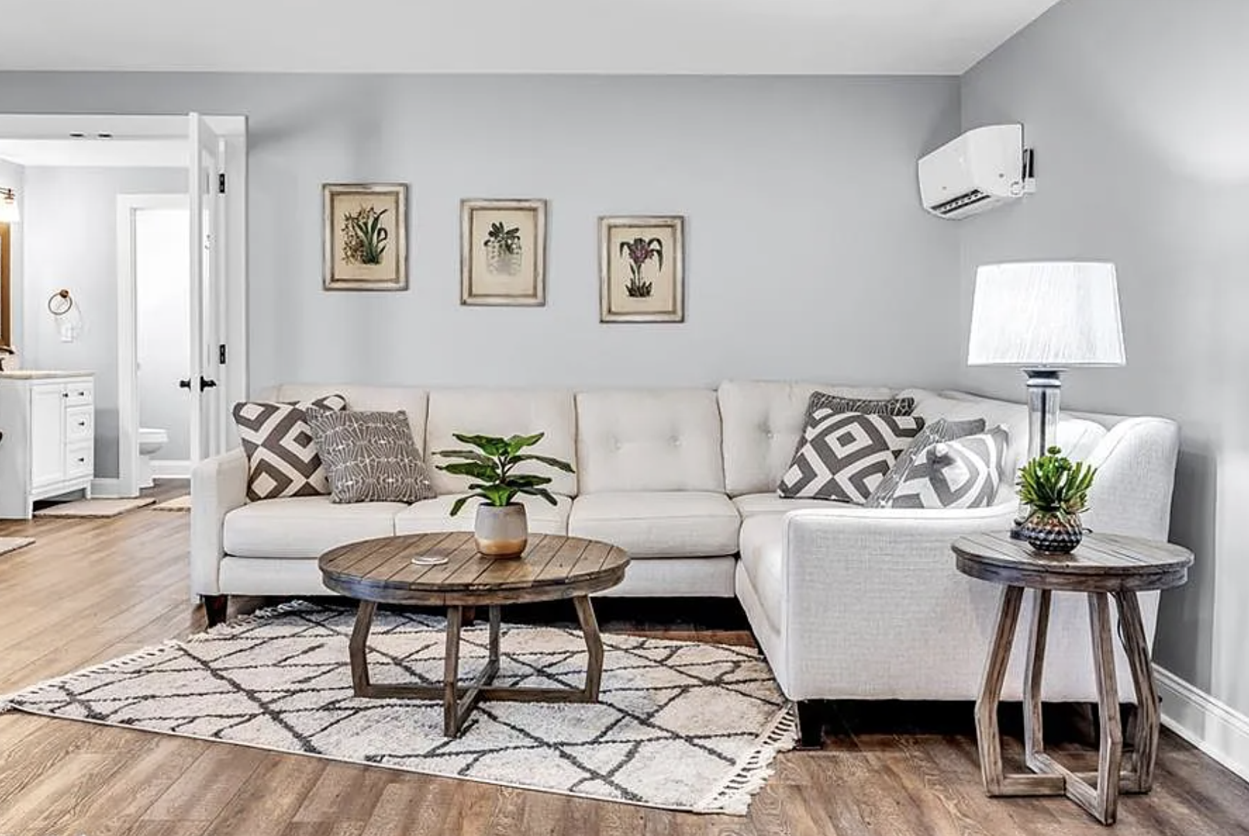 Living room with a white sectional sofa, patterned cushions, wooden coffee table, side table with a table lamp, framed plant art on the wall, and an air conditioner.