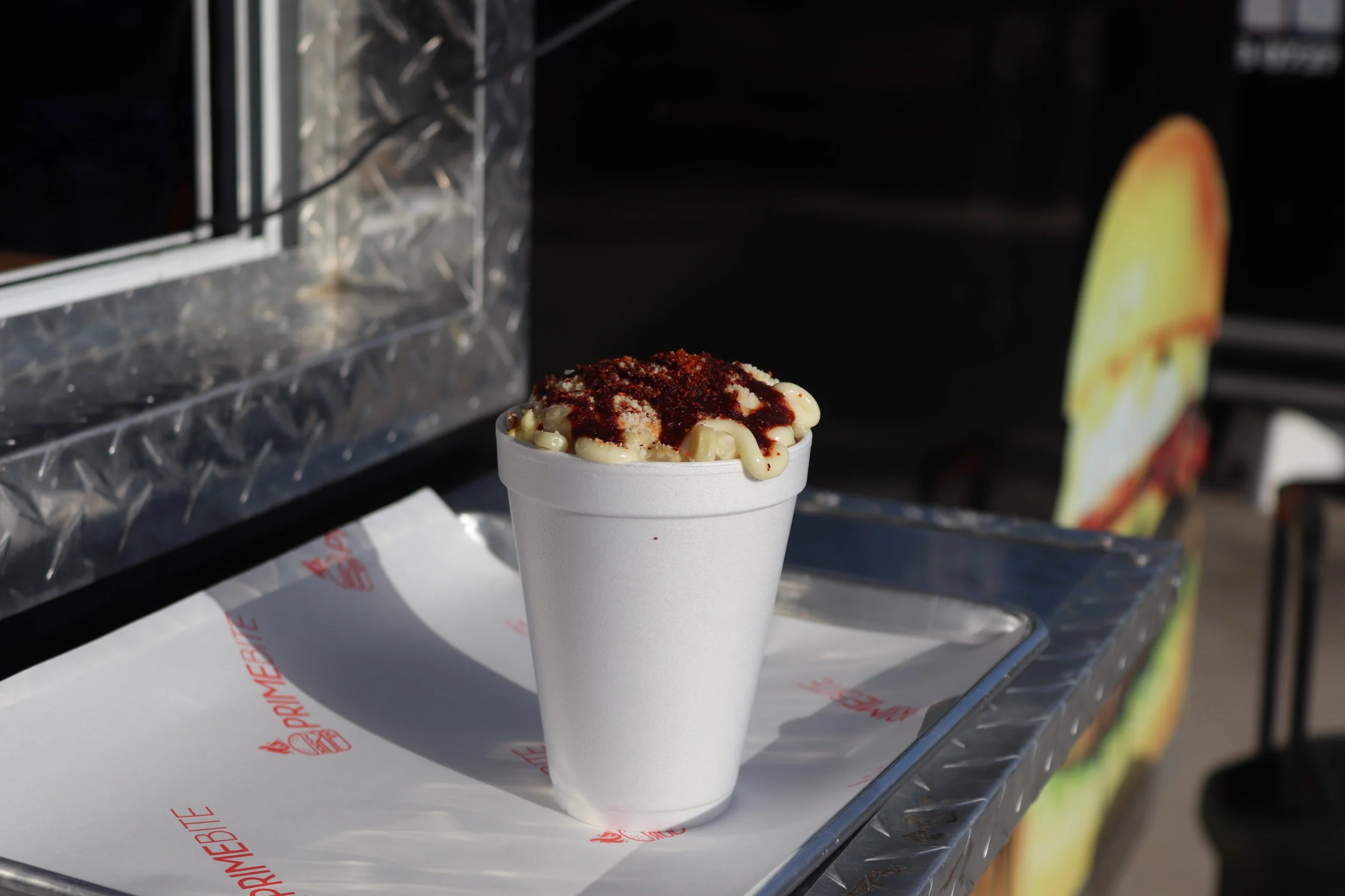 A cup of ice cream with chocolate syrup and a red sprinkle topping on a white foam cup, placed on a reflective metal surface with a black background.