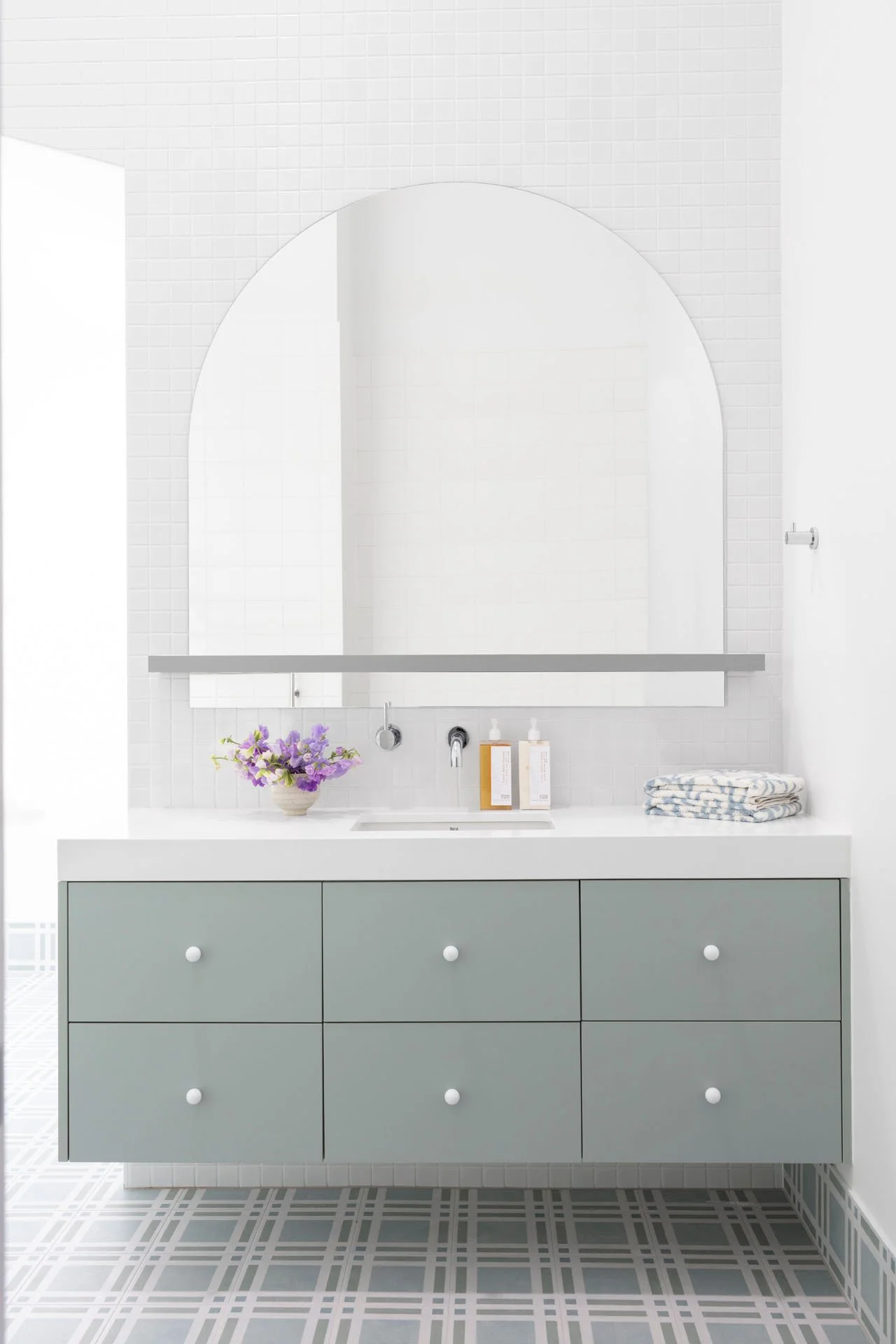 A modern bathroom vanity with a large mirror, six drawers, a white countertop, a small flower bouquet, and folded towels. The wall behind is tiled white, and the floor has a geometric-patterned tile.