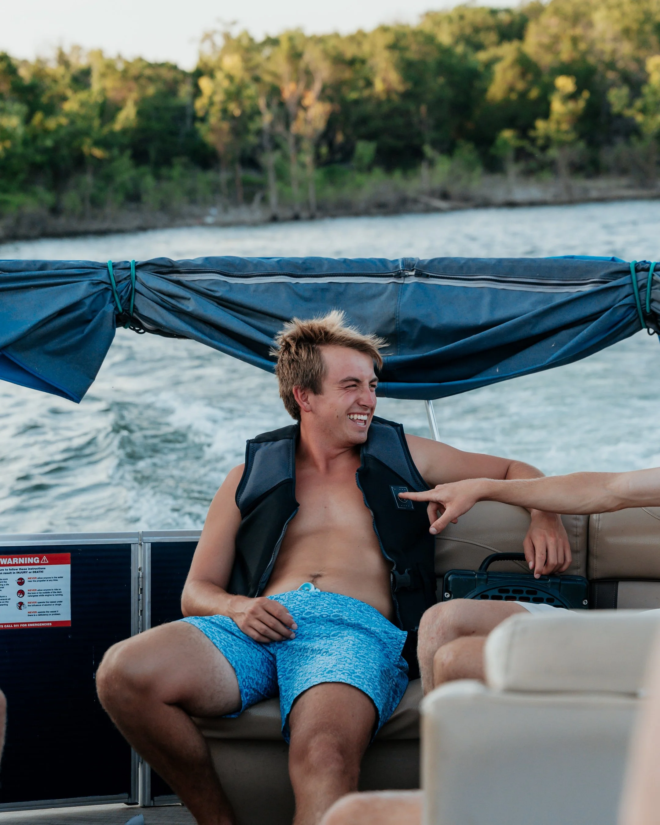 A young man in a life vest and blue swim trunks sitting on a boat, smiling and laughing as someone points a finger at his chest. The boat is on water with a green tree-lined shoreline in the background.