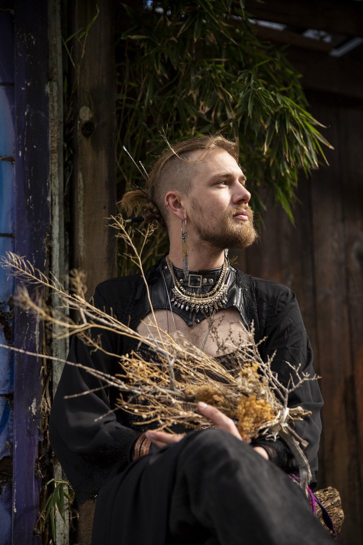 A man with a partially shaved head, long hair tied in a bun, and a beard, sitting outdoors with dry plants in his hands. He wears layered necklaces, earrings, and a black strappy top with a choker. Background includes wooden walls and green foliage.