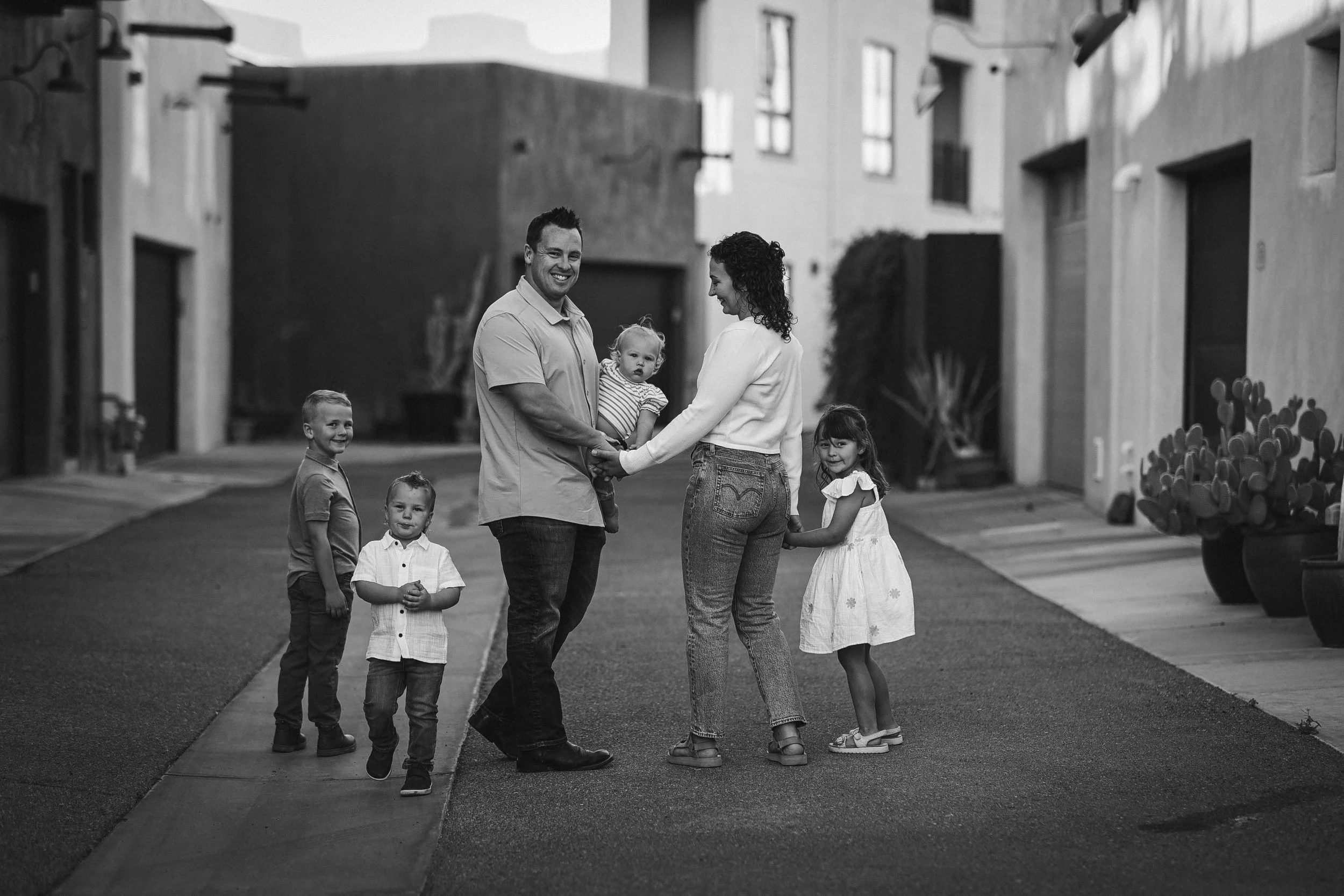 A family of six standing in the middle of a residential alleyway, holding hands and smiling. Four children and two adults, with the adults in the center and children surrounding them.