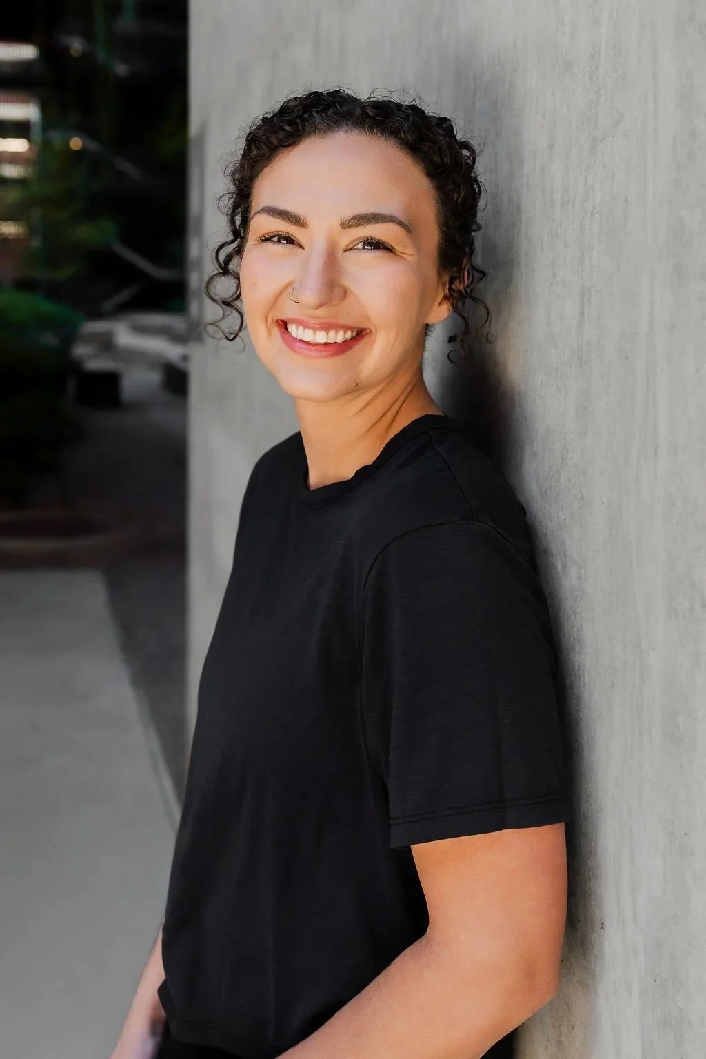 A woman with short, curly dark hair, smiling and wearing a black shirt, leaning against a concrete wall outdoors.