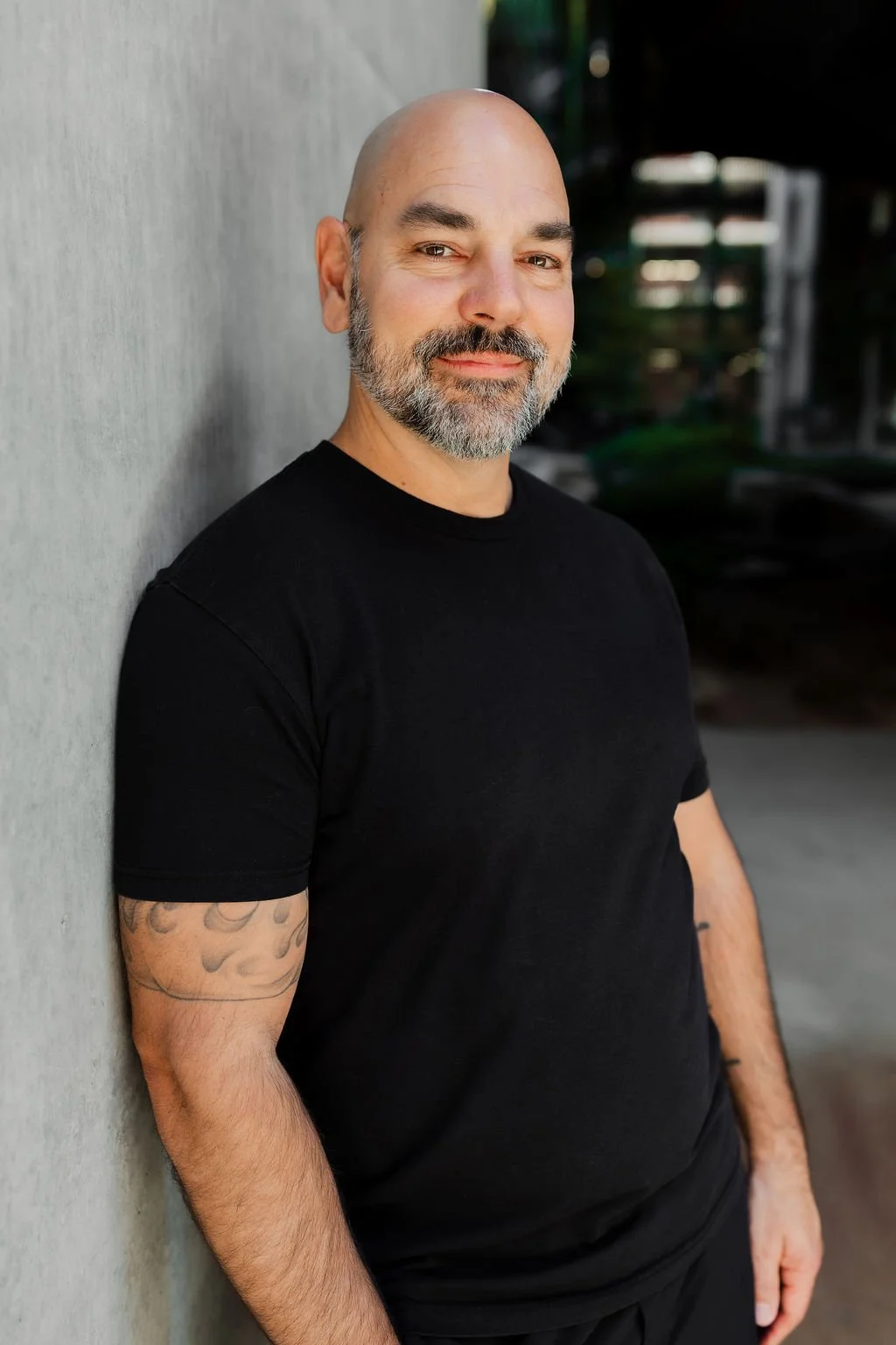 A bald man with a salt and pepper beard, wearing a black T-shirt, stands leaning against a gray concrete wall, with a slight smile and relaxed posture, outdoor setting with blurred greenery in background.