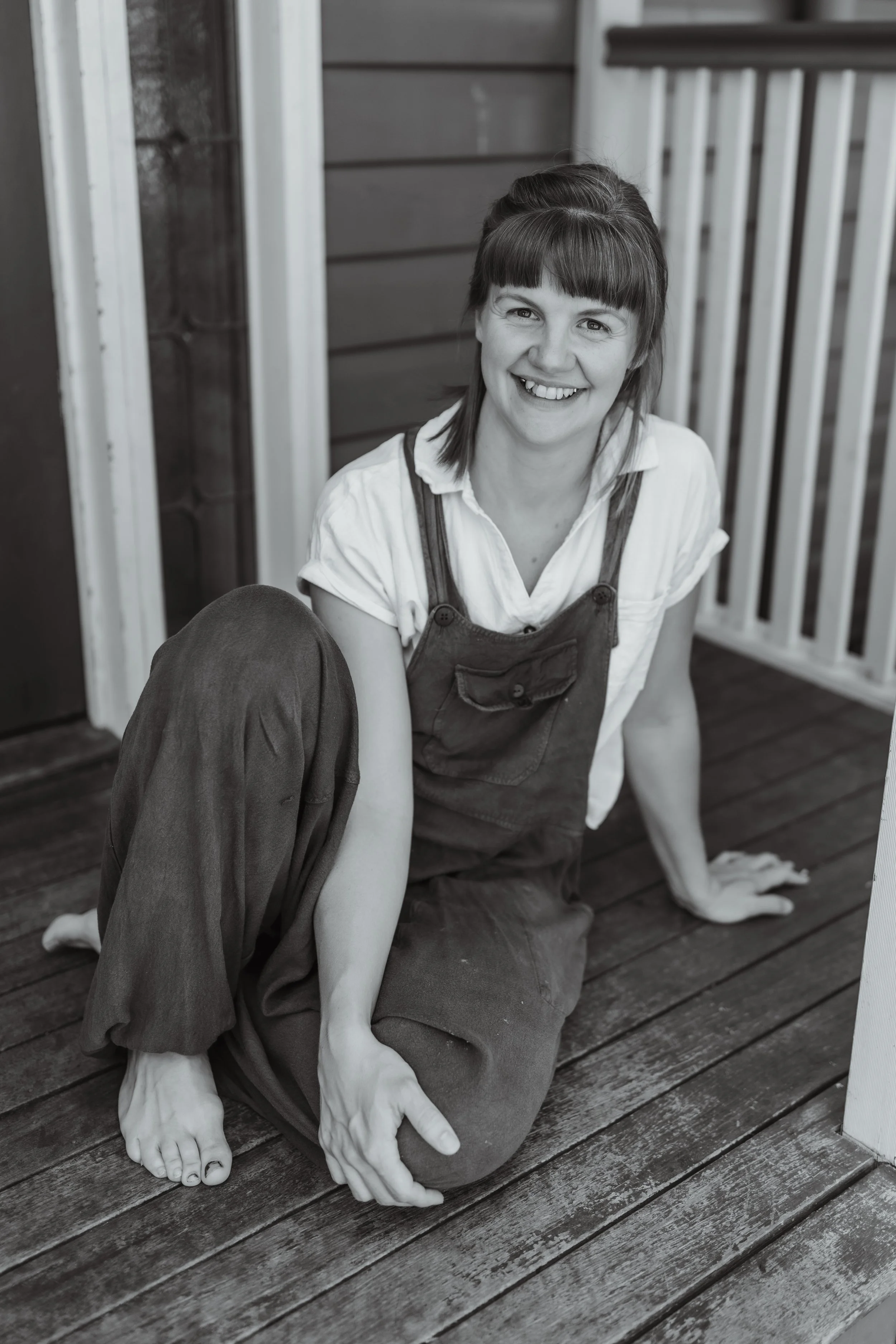 A woman smiling and sitting on a wooden porch, wearing overalls and a white shirt.