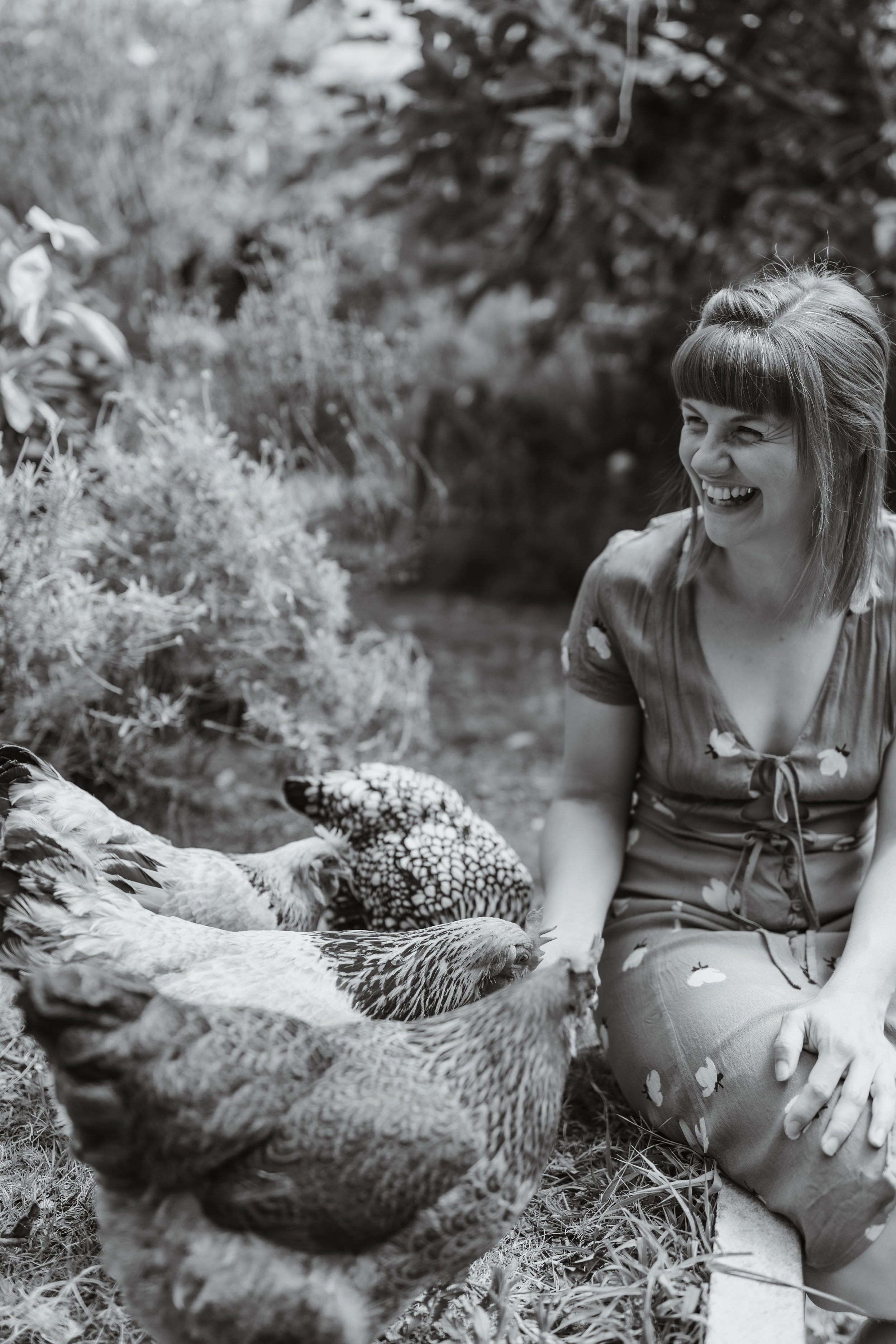 A woman sitting outdoors laughing with chickens around her.