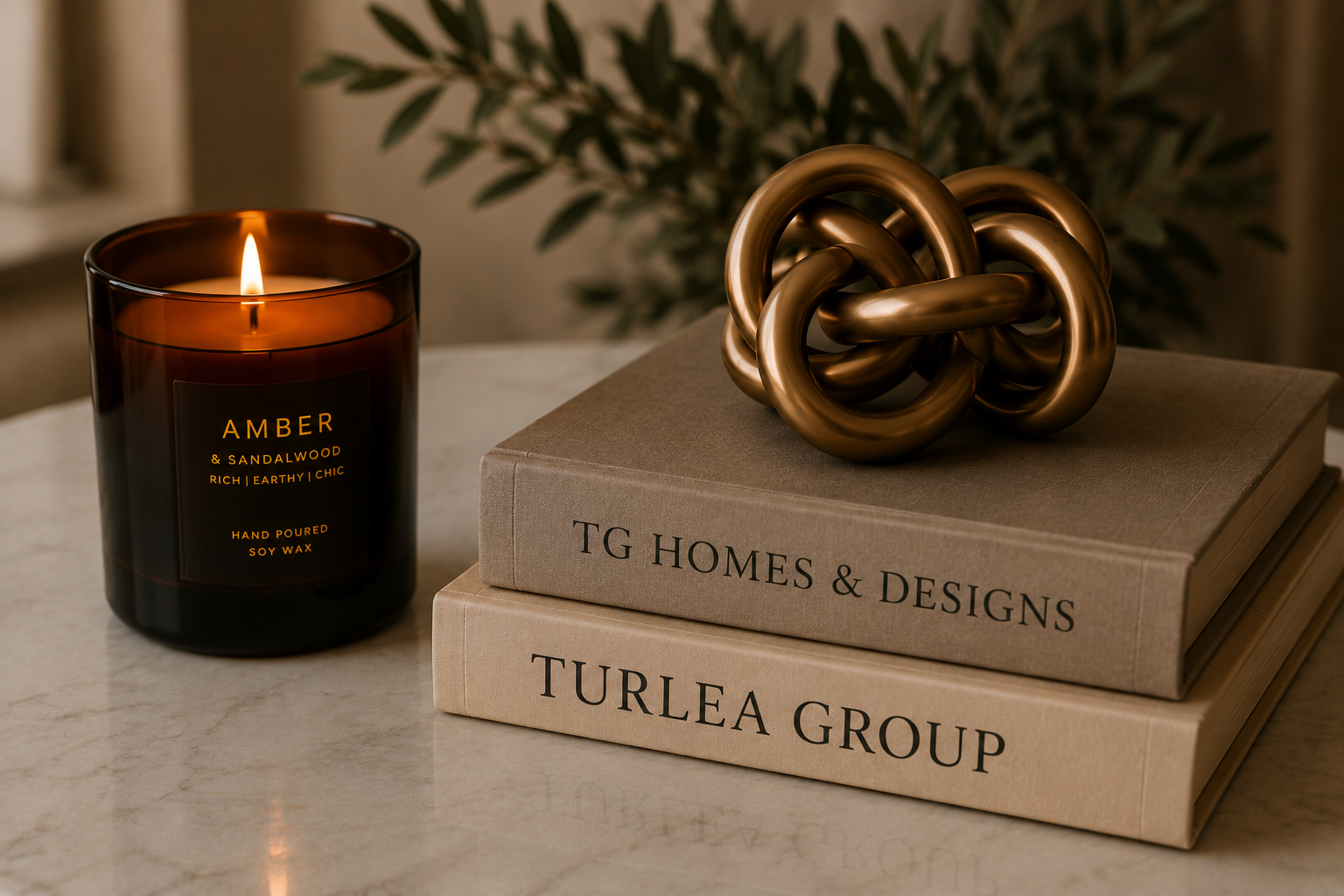 A lit amber candle with a label that reads 'Amber & Sandalwood, rich, earthy, chic, hand poured soy wax,' sits next to stacked books titled 'TG Homes & Designs' and 'Turlea Group.' on a marble surface, with a gold decorative knot on top and a blurred plant in the background.