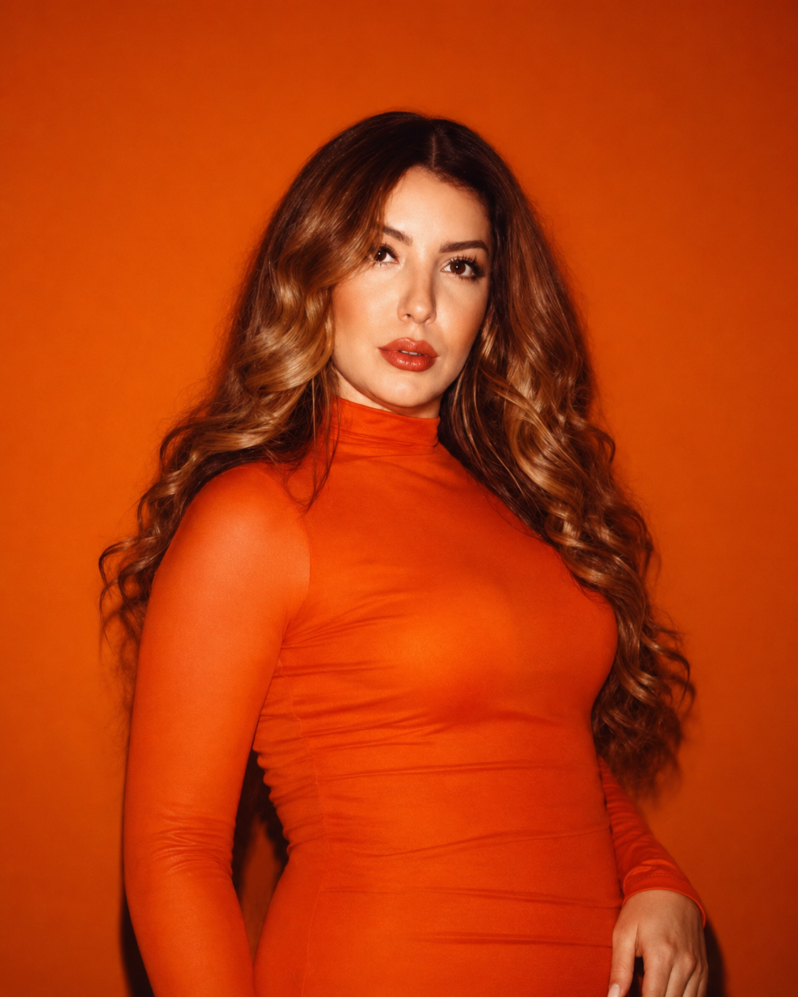 A woman with long, wavy brown hair wearing a bright orange, form-fitting long-sleeve top, standing against an orange background.