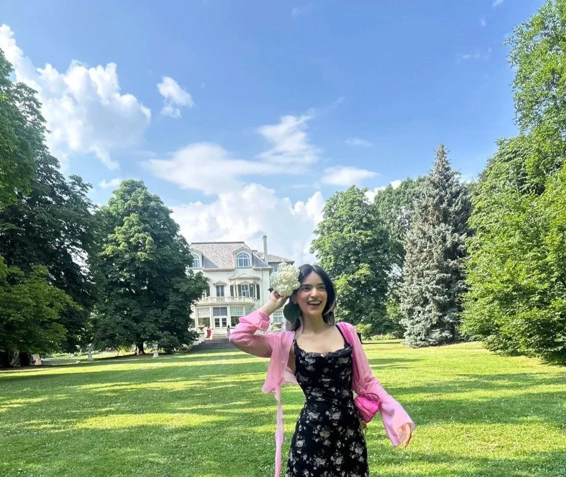 A woman standing on a grassy area in front of a large house, holding a bouquet of white flowers to her head, smiling, wearing a black floral dress and a pink jacket, surrounded by green trees on a sunny day.