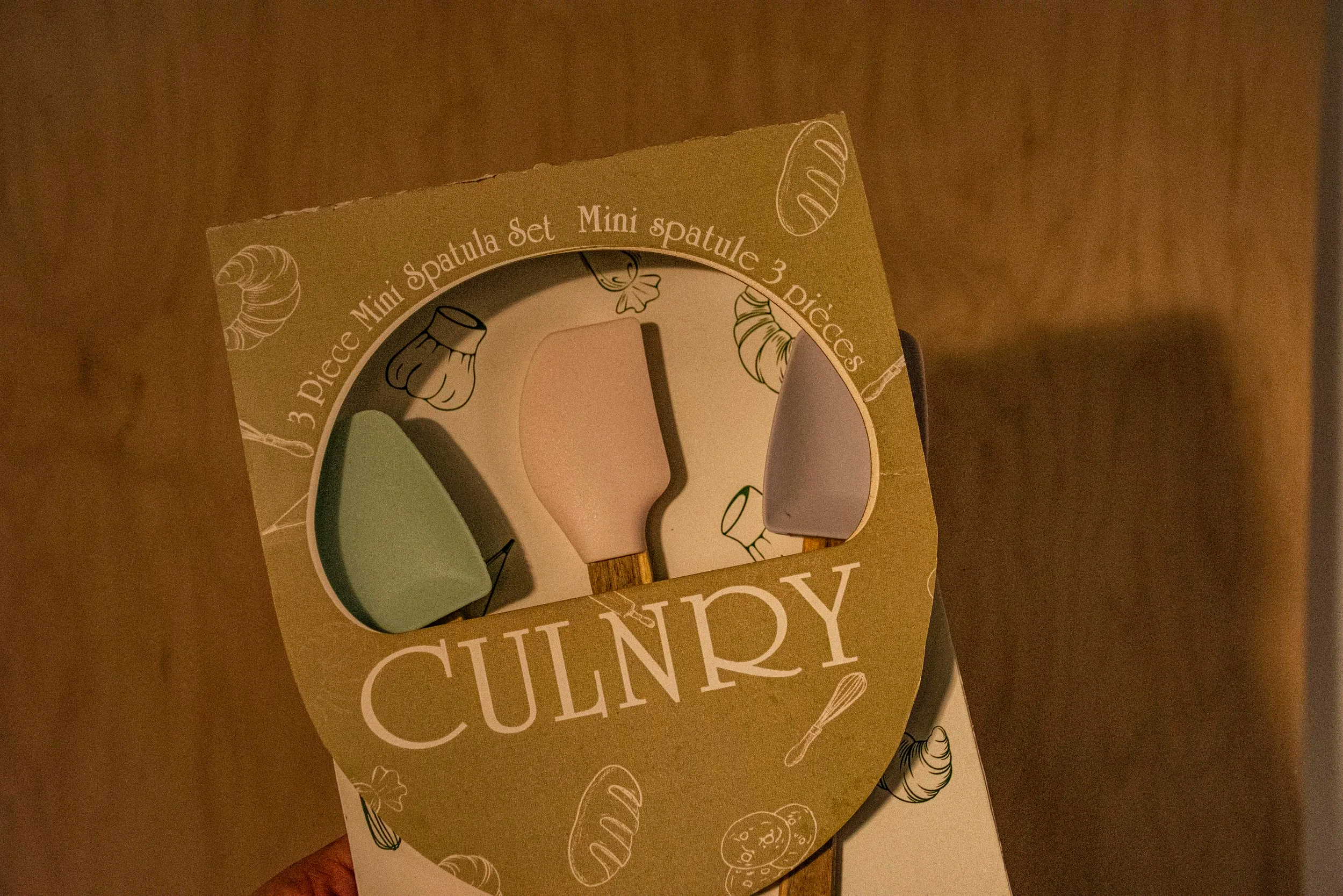 A hand holding a kitchen utensil set in a cardboard box with three pastel-colored spatulas, labeled as a "Mini Spatula Set" with three pieces.