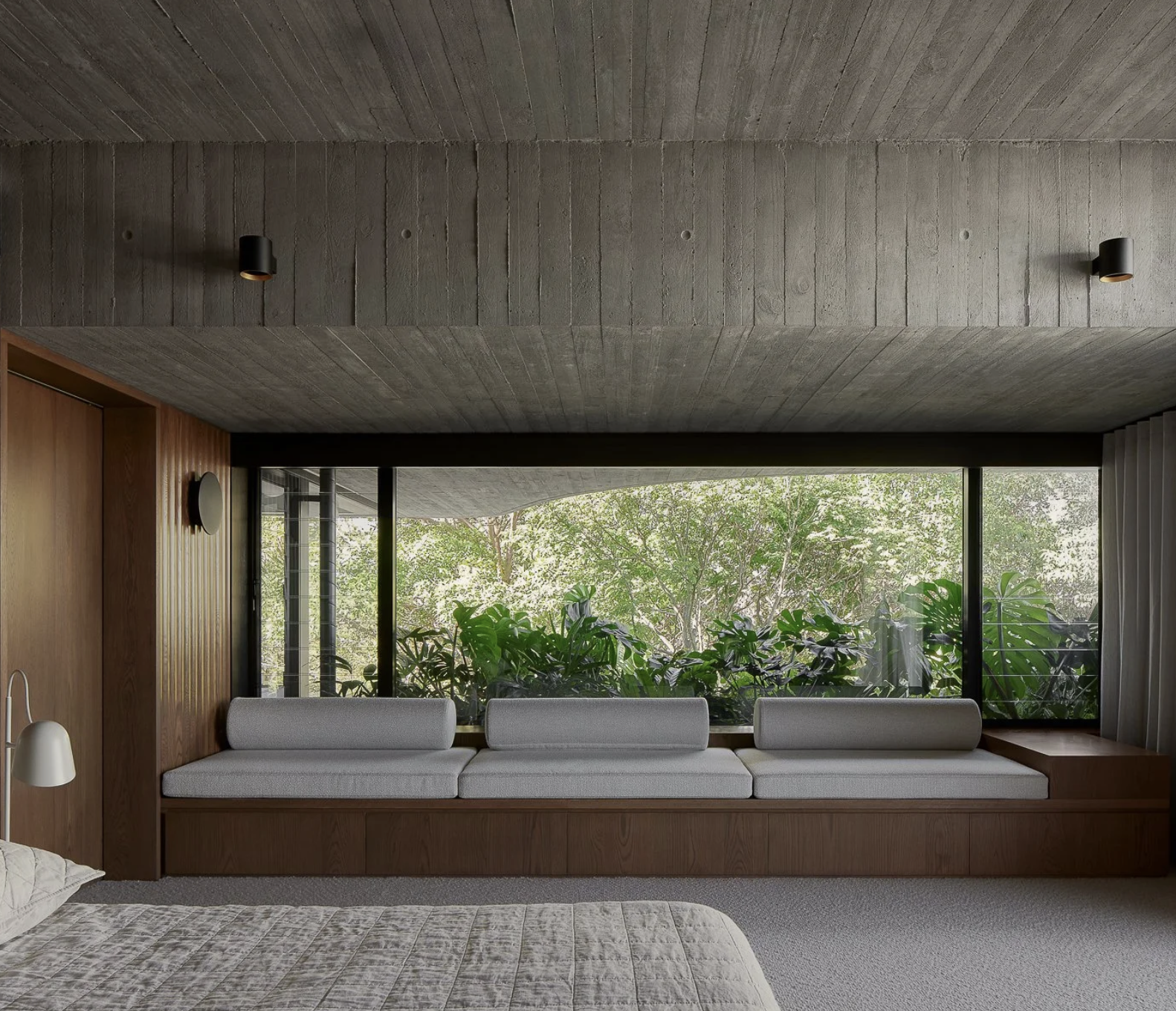 Modern hotel room with large window seating area and greenery outside, gray ceiling and wooden wall with small wall light.