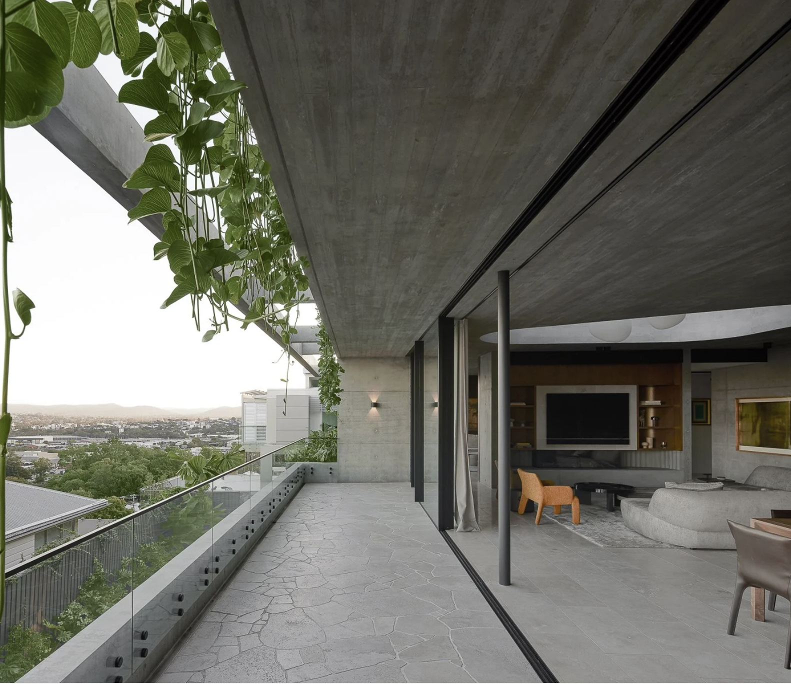 Modern apartment balcony with green vines hanging from the ceiling, overlooking a cityscape with trees and buildings in the distance, and visible interior living room through glass sliding doors.