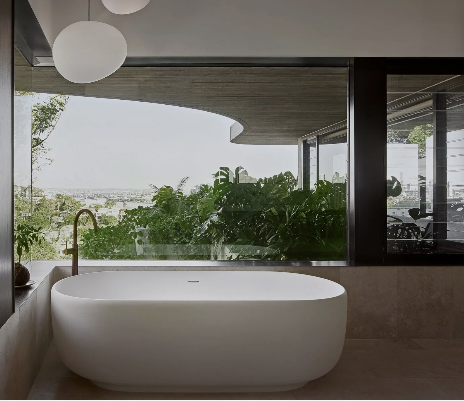 Modern freestanding bathtub with a view of lush green plants and cityscape through large windows.