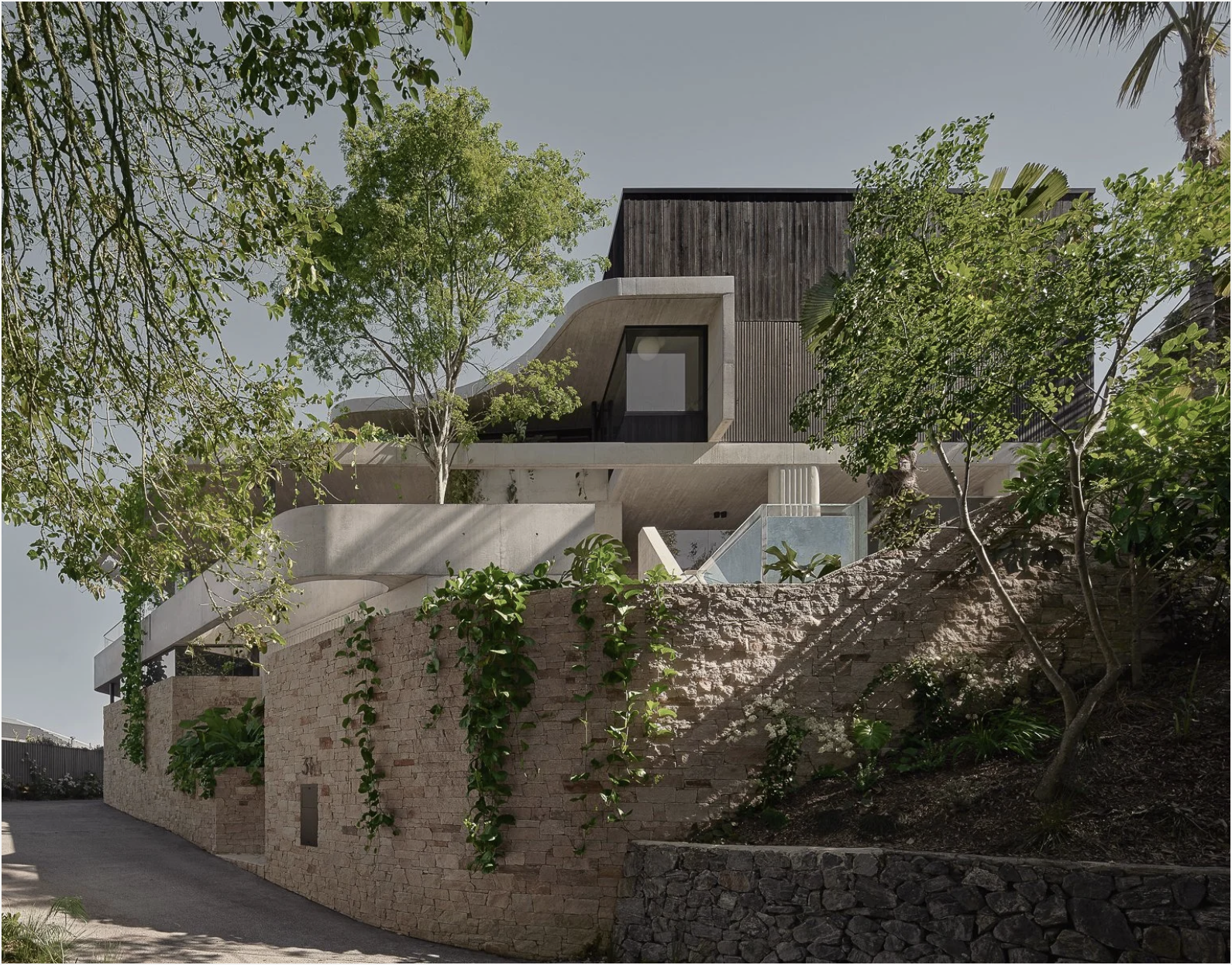 Modern multi-level house with wooden and concrete exterior surrounded by trees and greenery, situated on a sloped terrain with stone retaining walls.