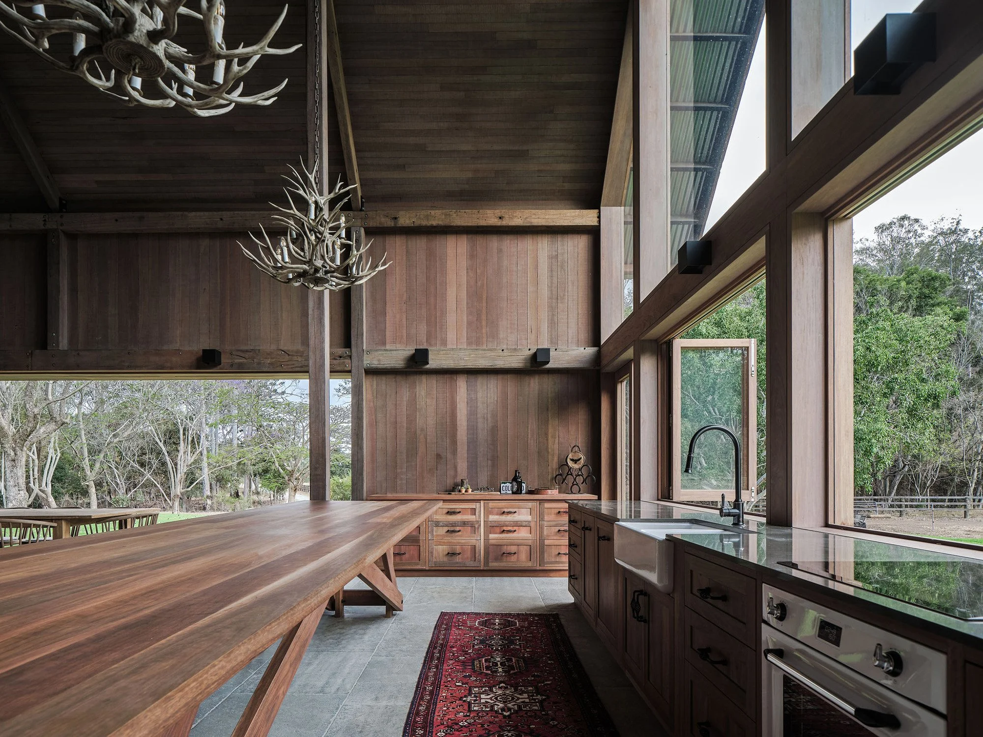 Interior view of a rustic kitchen with large wooden dining table, wooden cabinets, a farmhouse sink, and large windows showing a wooded outdoor scene, with antler chandeliers hanging from the ceiling.
