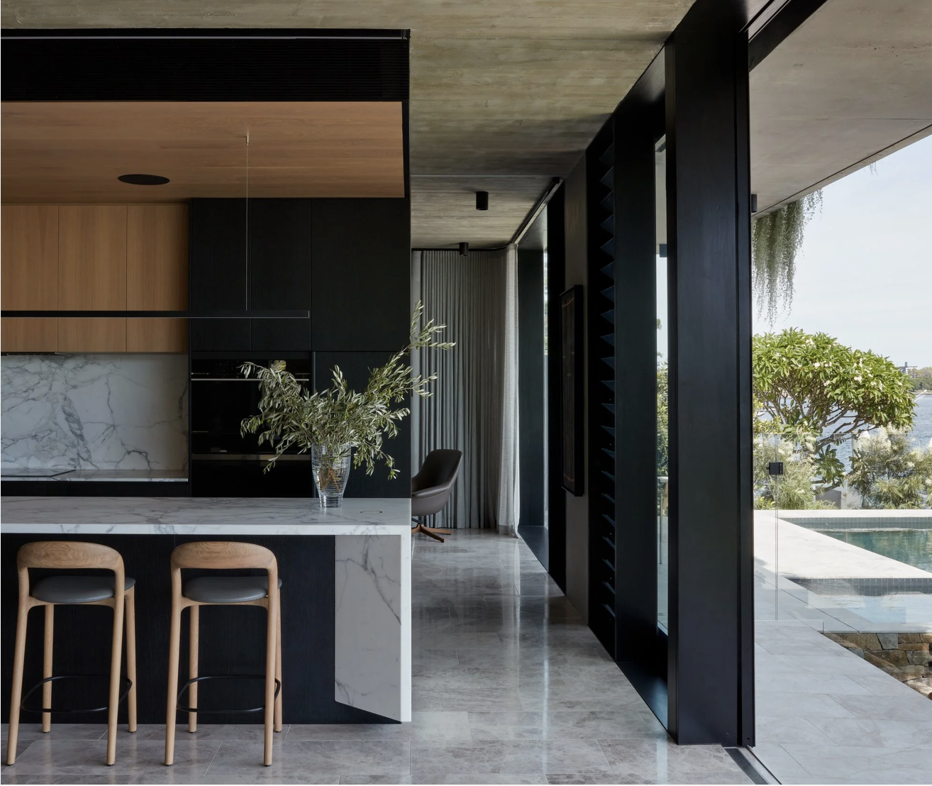 Modern kitchen with black and wood cabinetry, marble countertop, and an open view to a pool and greenery outside.