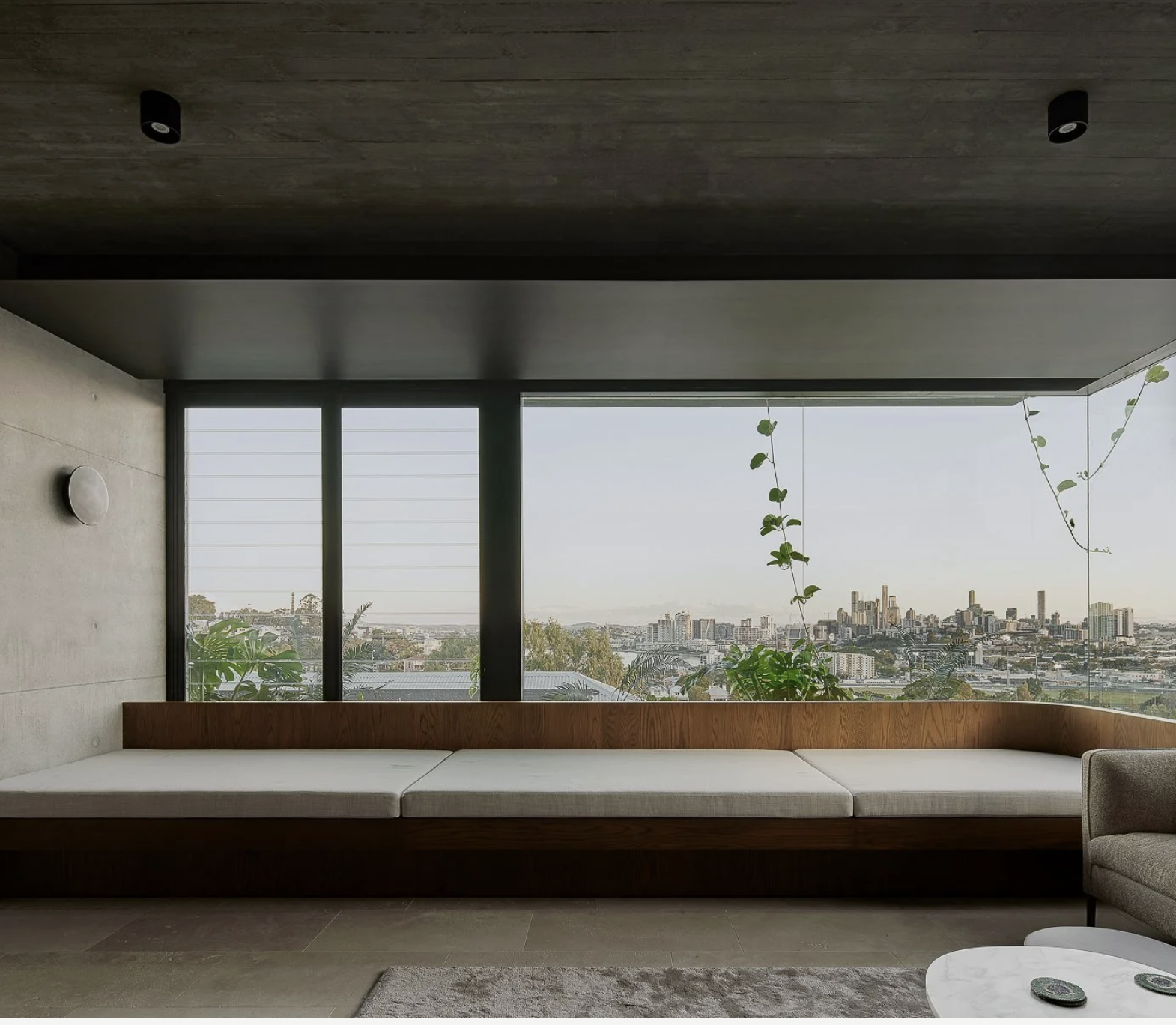 Modern interior with a long wooden bench with cushions, large windows showing city skyline, plants hanging from the ceiling, and a small round table with remote controls.