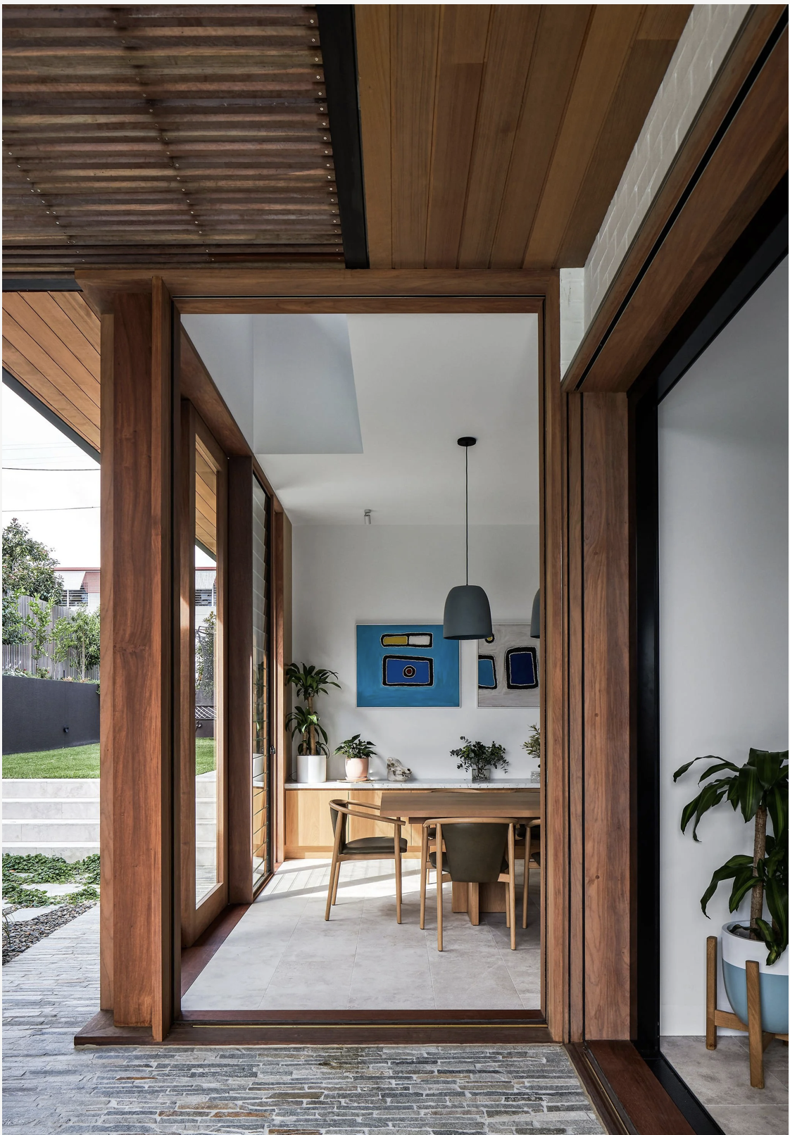 Modern dining room with wooden accents, white walls, and plants, visible through a doorway.