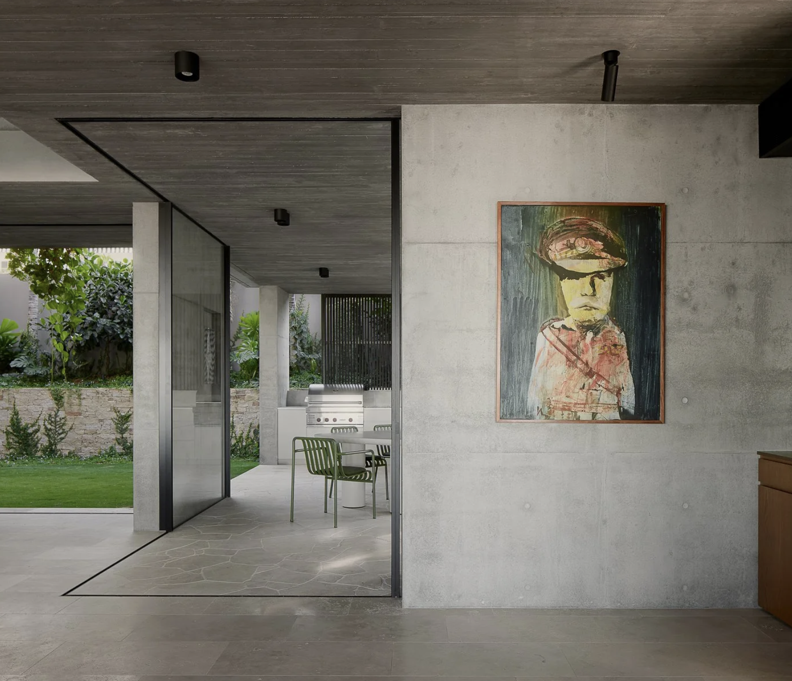 Modern interior with concrete walls and ceiling, a colorful abstract portrait of a young boy wearing a cap hanging on the wall, and a view into an outdoor patio with green chairs, a barbecue grill, and lush garden greenery.