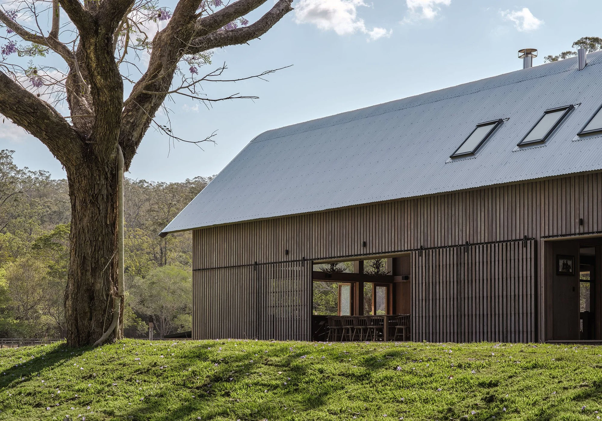 A modern barn with a metal roof, wood siding, and skylights surrounded by green grass and trees in a rural setting.