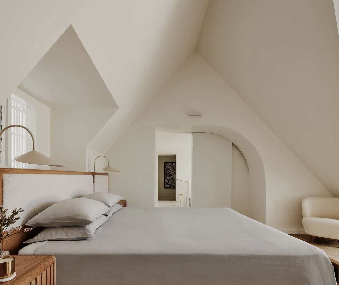 Bedroom with white walls and sloped ceiling, bed with gray pillows and wooden headboard, white side table with plant, wall-mounted lamps, and a cream armchair