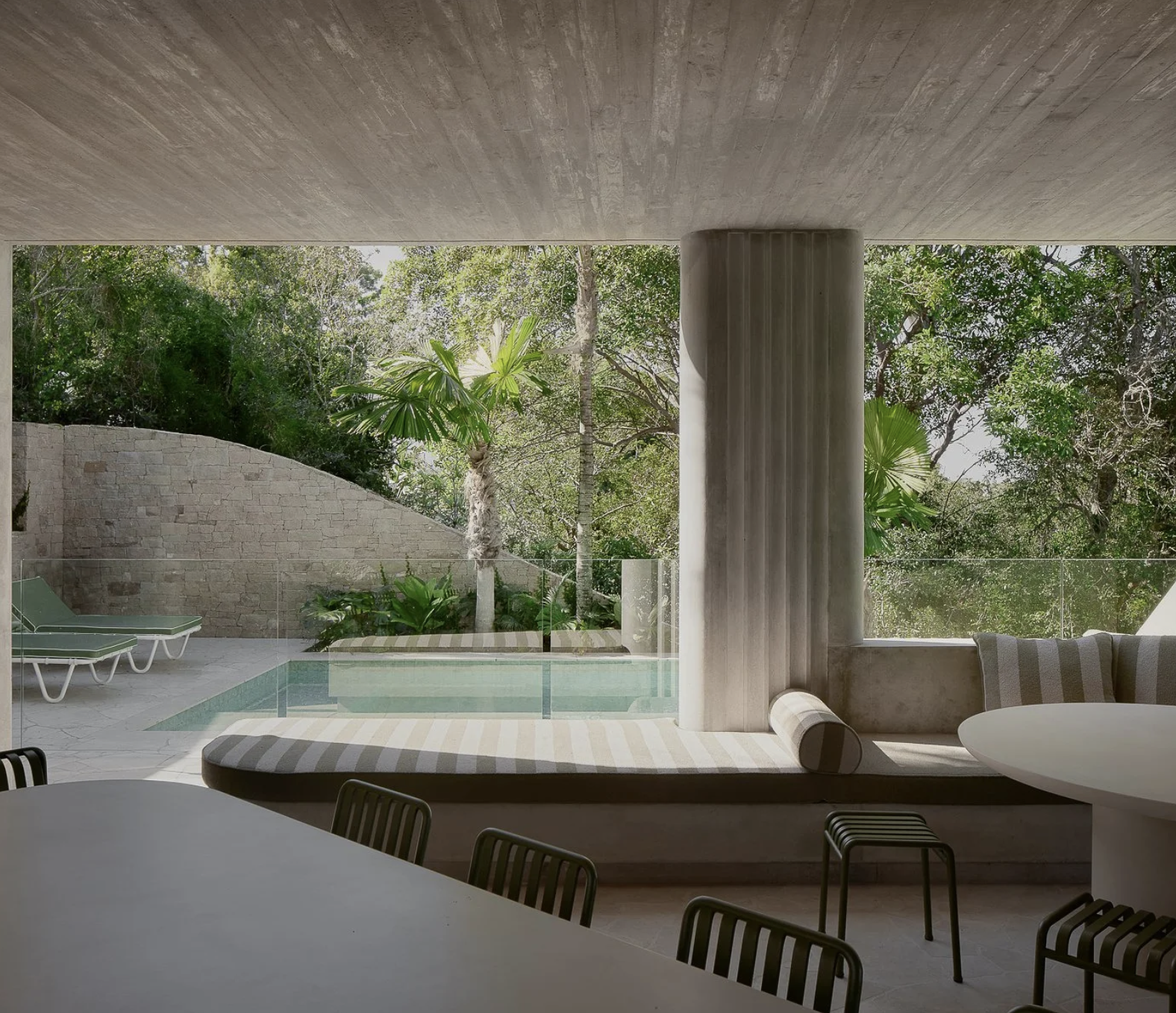Modern indoor space with a view of a pool and lush greenery outside, featuring a built-in seating area with cushions, umbrellas, and a concrete pillar.