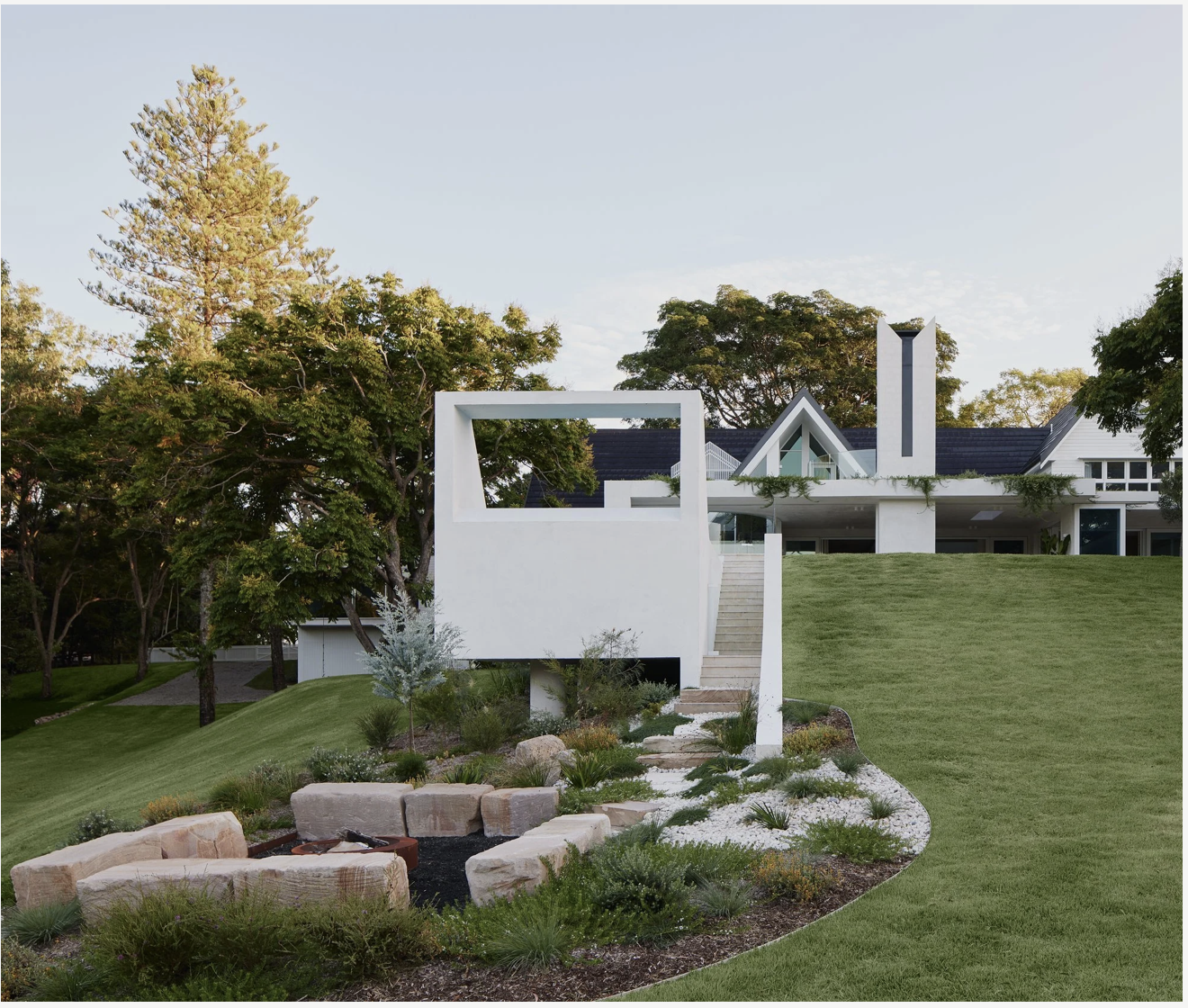 Modern house with white exterior, stairs leading to the entrance, surrounded by a landscaped yard with rocks and plants, tall trees in the background, and a clear sky.