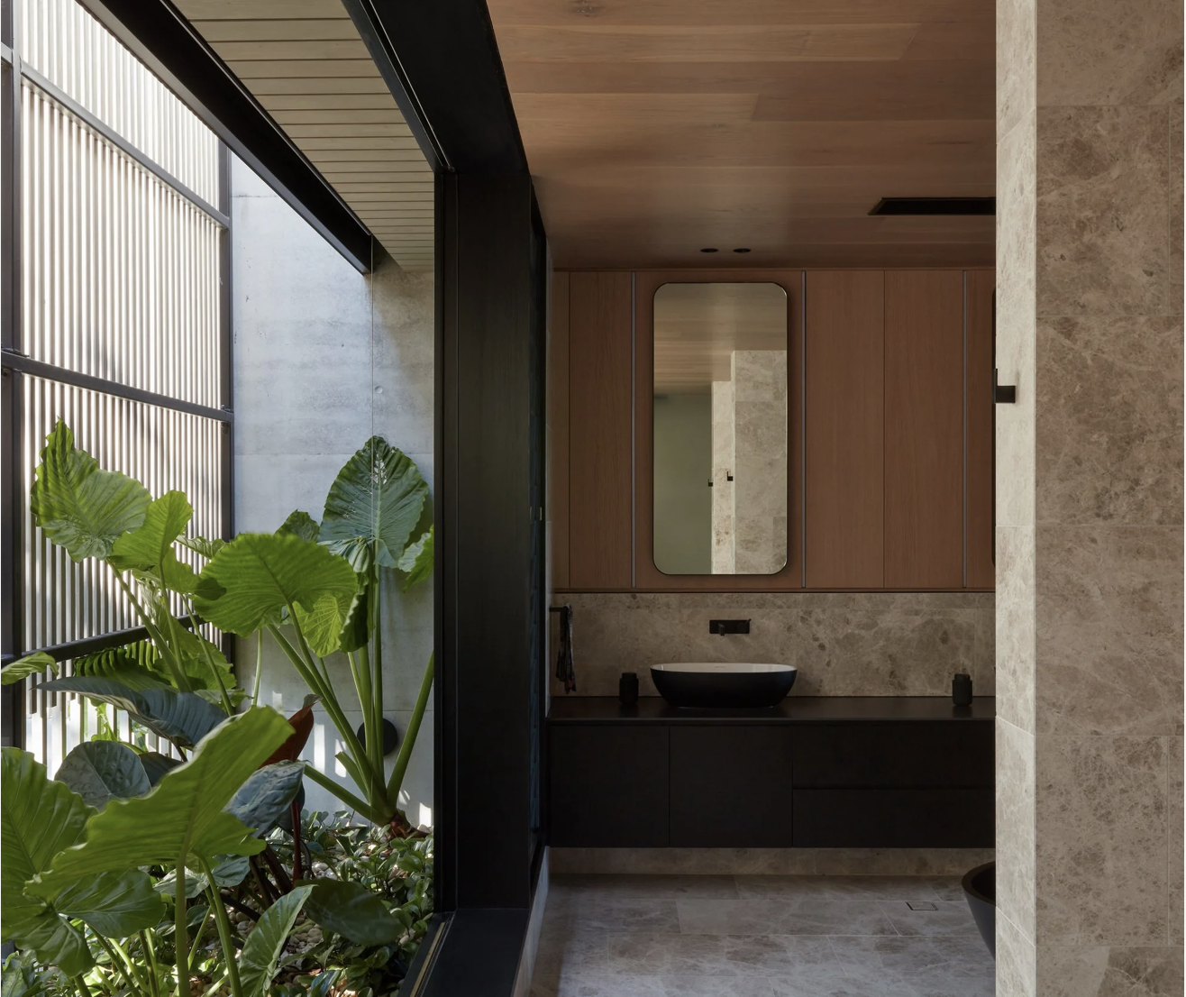 Modern bathroom with natural light, featuring a large window with plants outside, wooden cabinets, a mirror, and a black sink on a beige stone countertop.