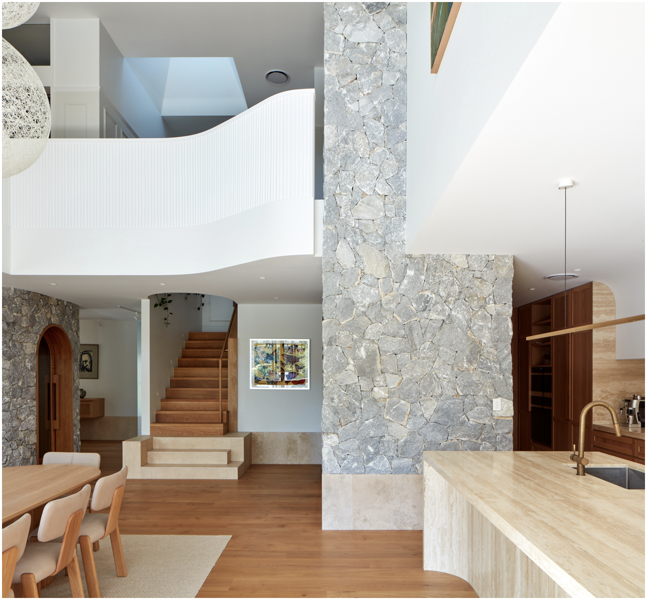 Interior view of a modern home featuring a stone wall, wooden staircase, and open kitchen with a beige marble island and wooden cabinets.