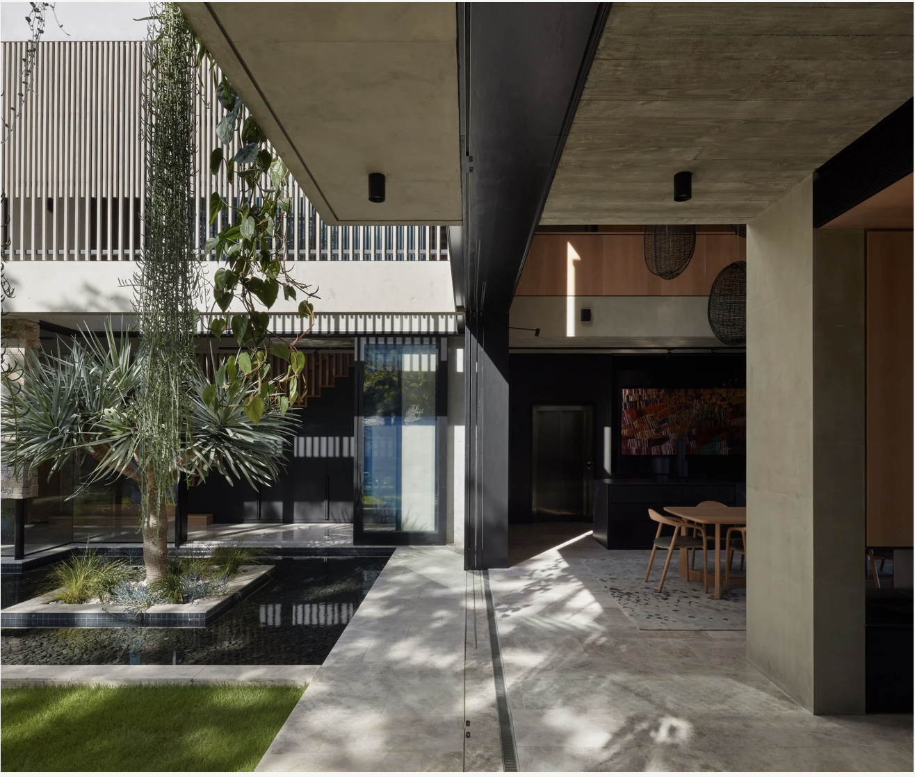 Modern interior courtyard with a raised garden bed, plants, and a water feature, visible through a glass door, with a dining area inside featuring wooden table and chairs, and decorative hanging lighting.