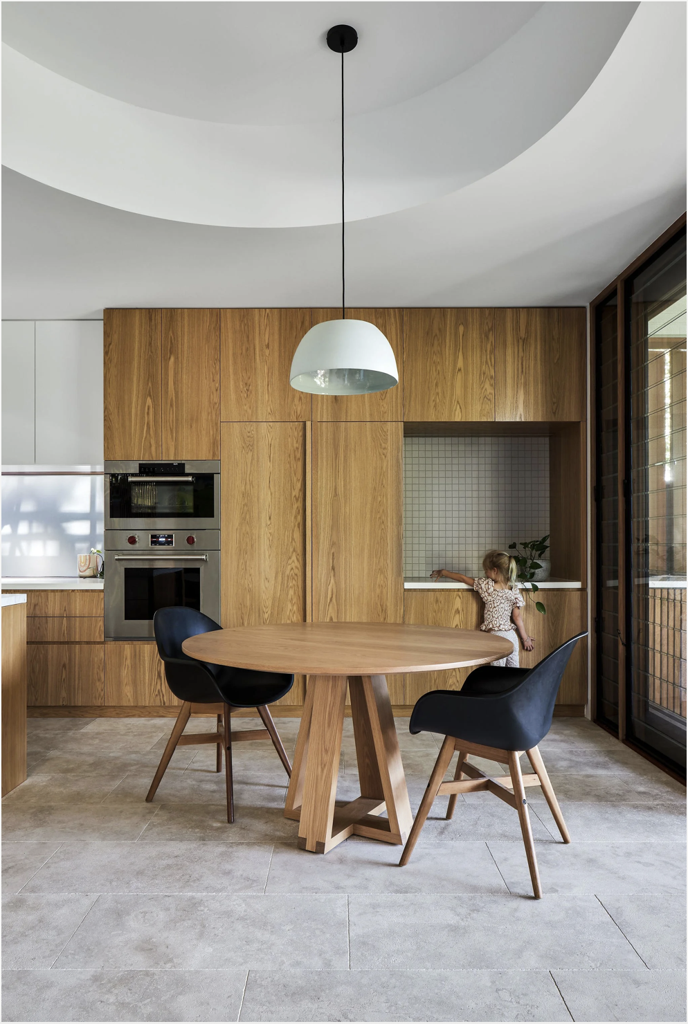 Modern kitchen with wooden cabinets, round wooden dining table, two black armchairs, a white hanging pendant light, child reaching for a potted plant near a window.