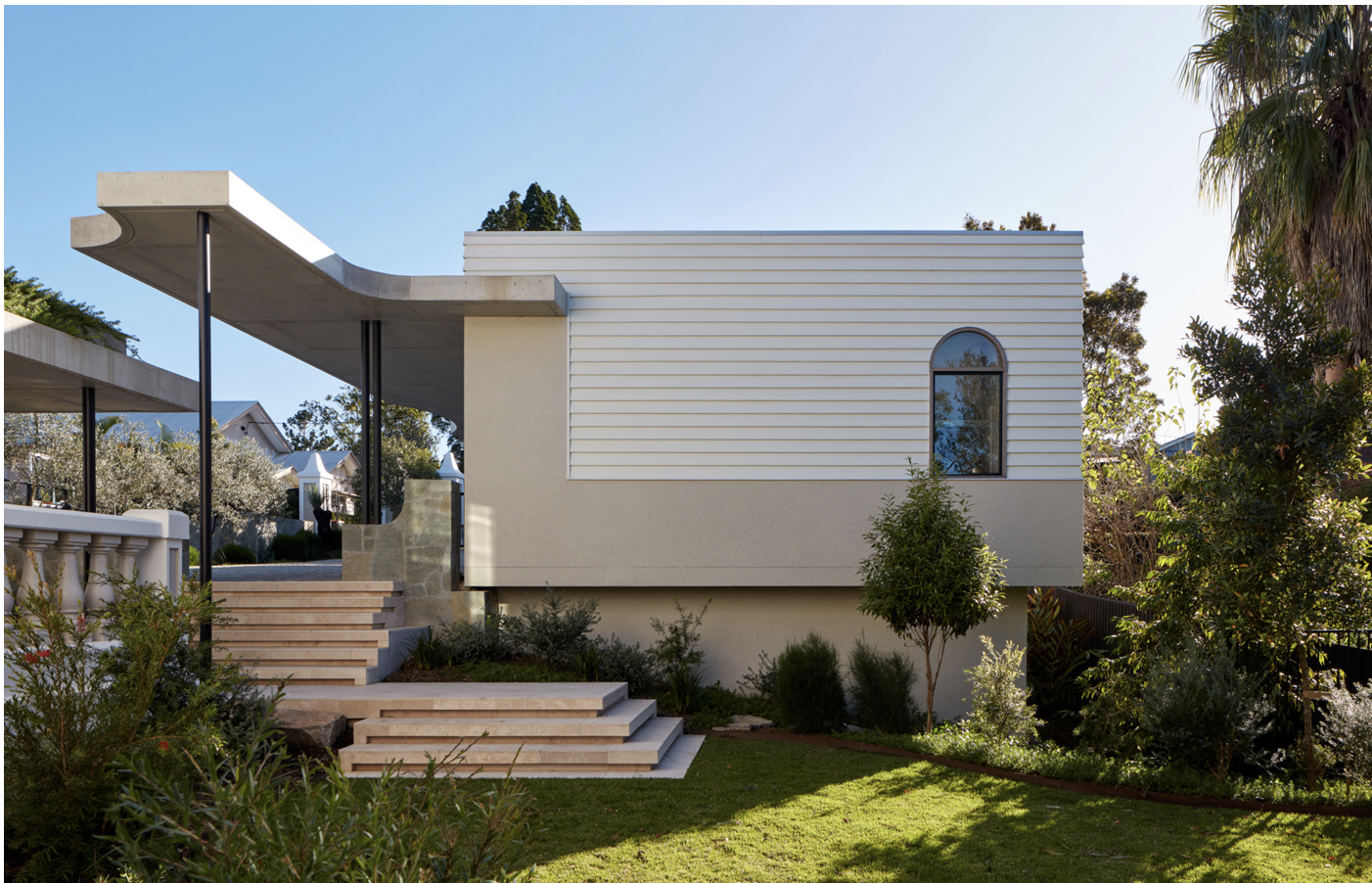 Modern house with white exterior, arched window, concrete stairs, and lush landscaped yard under clear blue sky.