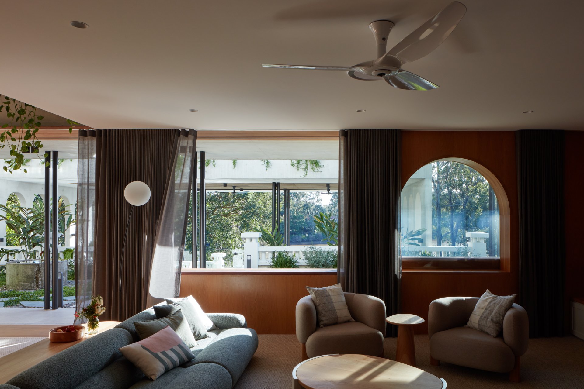 Living room with a ceiling fan, large windows, a gray sofa with throw pillows, and two beige armchairs with throw pillows, a small round wooden table, and curtains, with outdoor greenery visible through the windows.