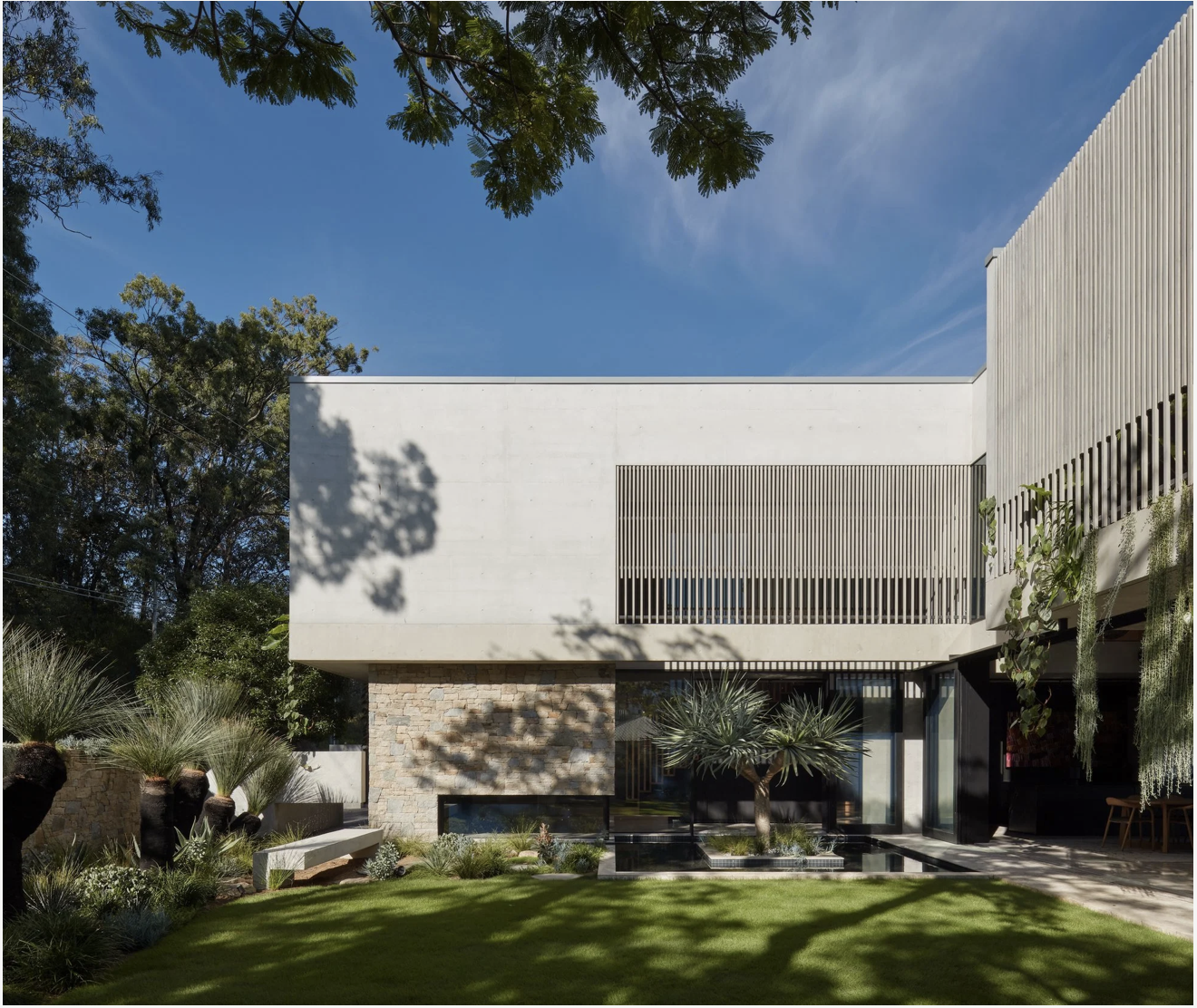 Modern house with white and stone facade, surrounded by landscaped garden with desert plants, trees, and shadows under blue sky.