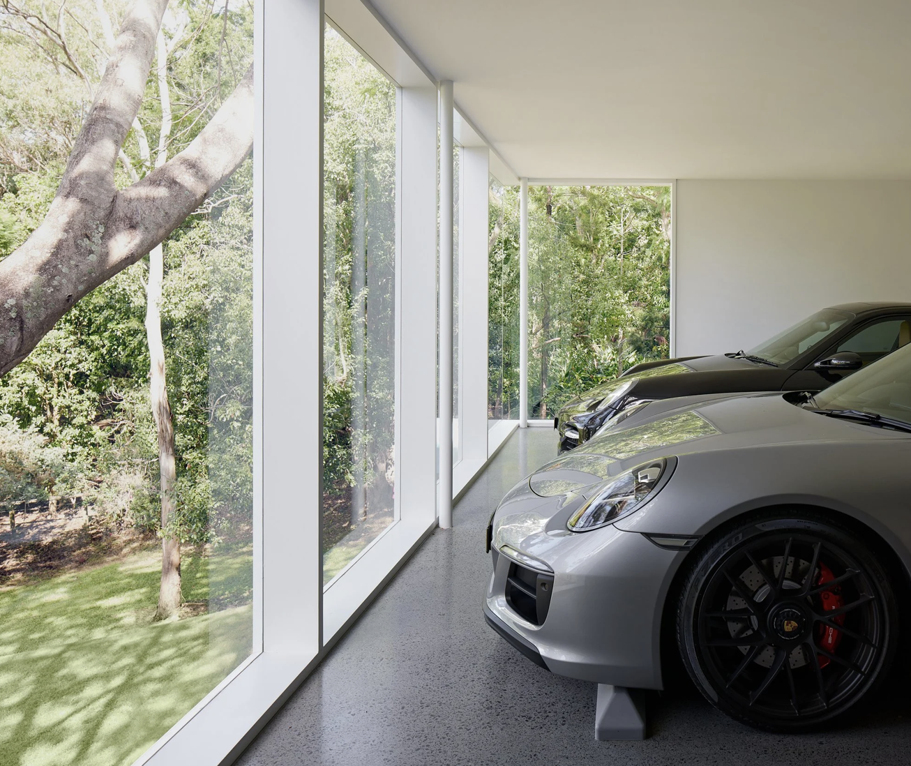 Modern garage with three sports cars parked inside, large floor-to-ceiling windows showing a view of trees and greenery outside