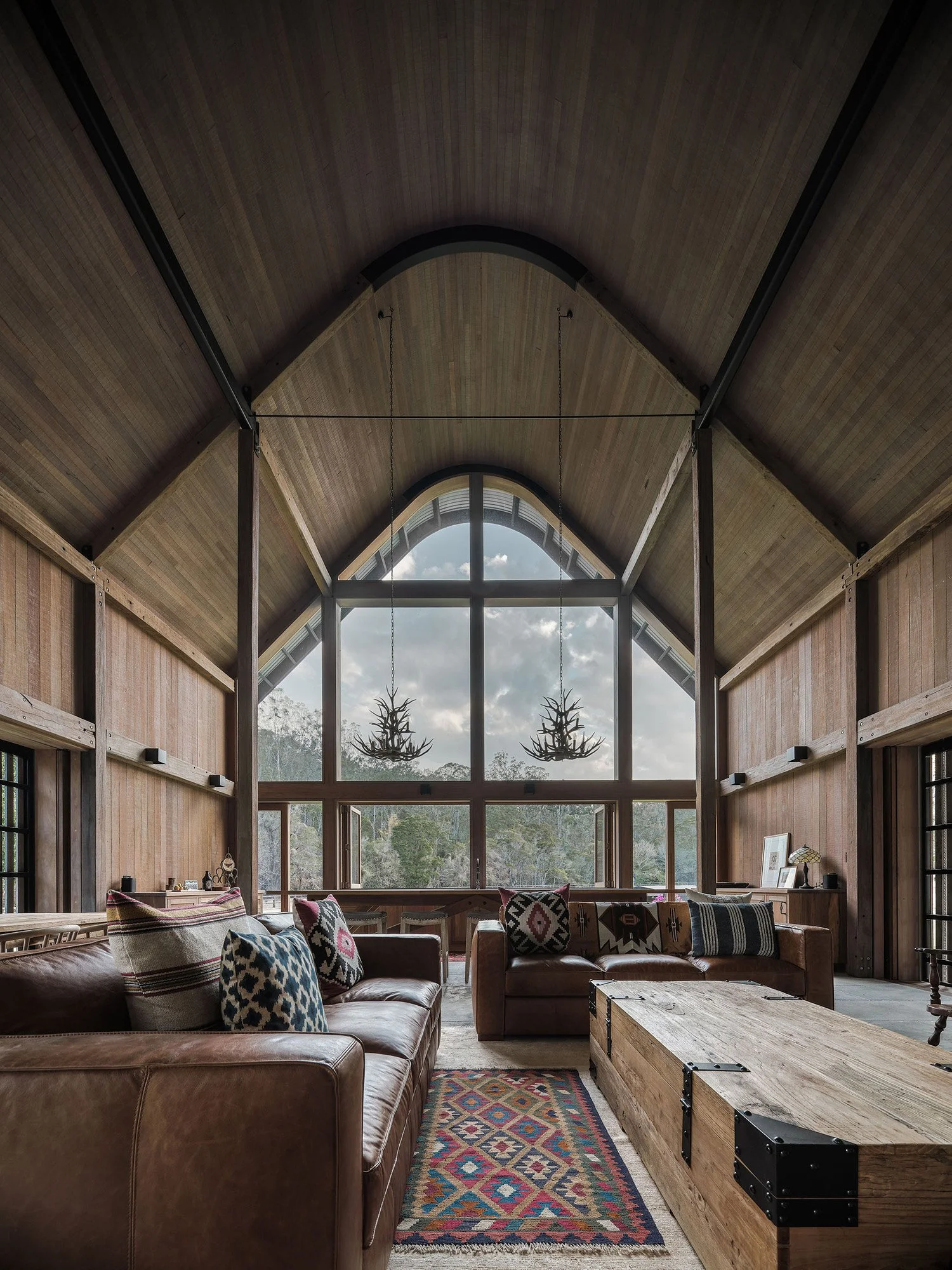Interior of a spacious living room with high vaulted wooden ceiling, large front window, hanging antler chandeliers, leather sofas with patterned pillows, a colorful rug, and a rustic wooden coffee table.
