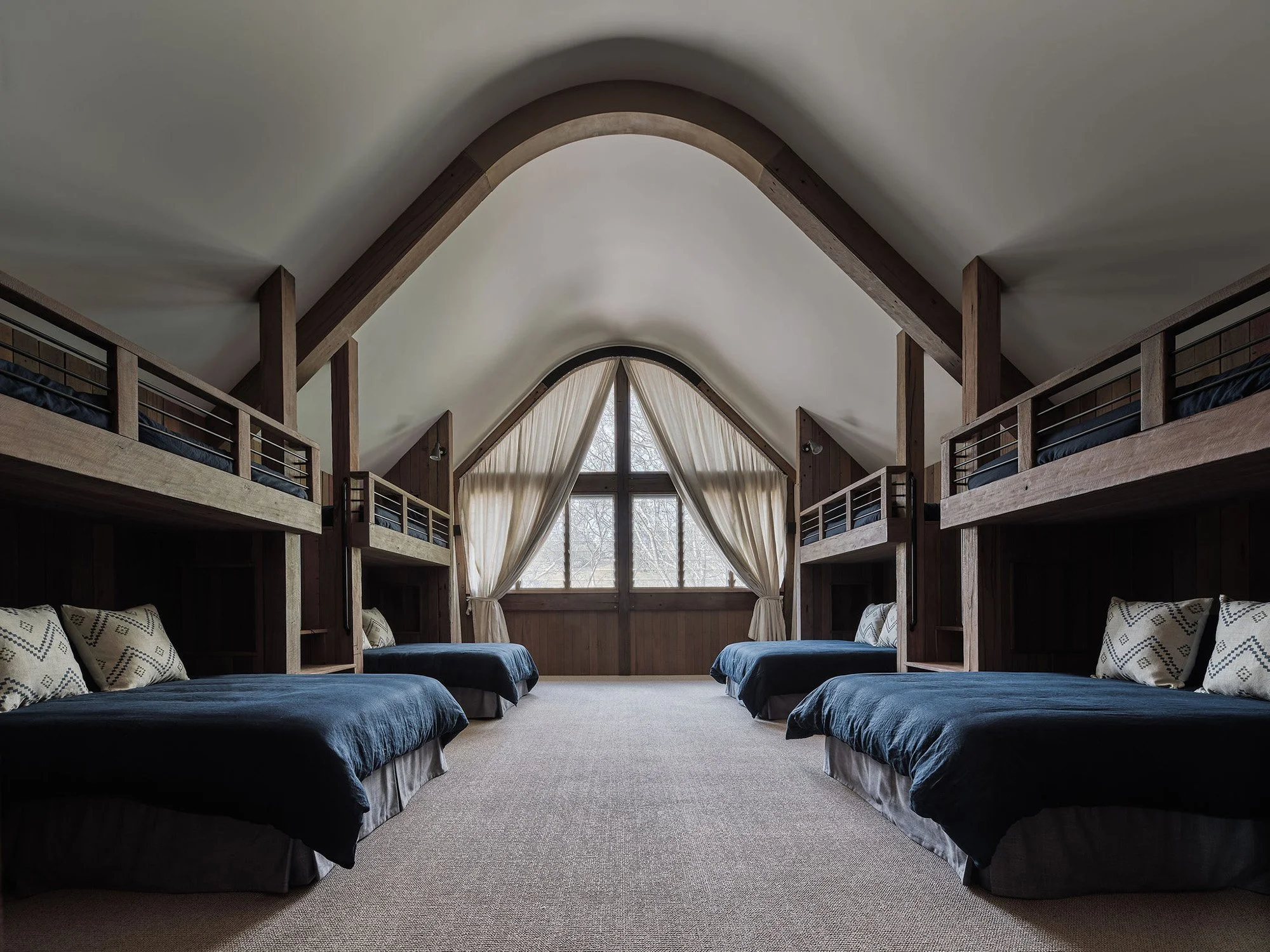 Room with four sets of twin beds, each with black bedding and patterned pillows, in a vaulted ceiling space with large window covered by beige curtains.