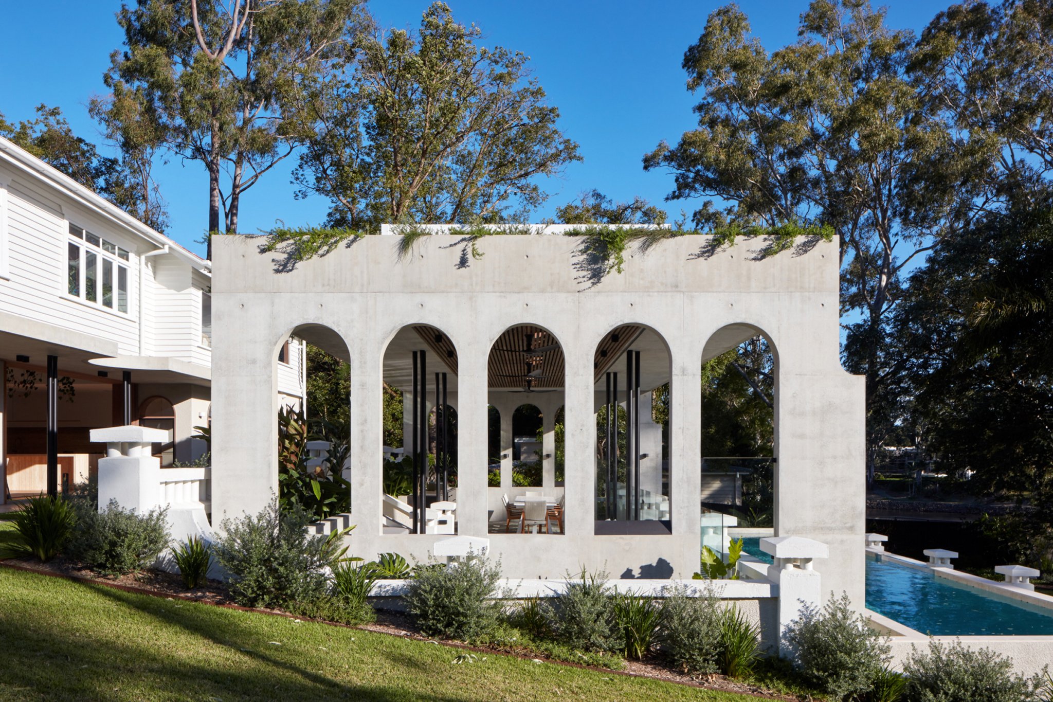 Modern outdoor structure with large arches and glass panels, situated next to a swimming pool, surrounded by greenery and trees.