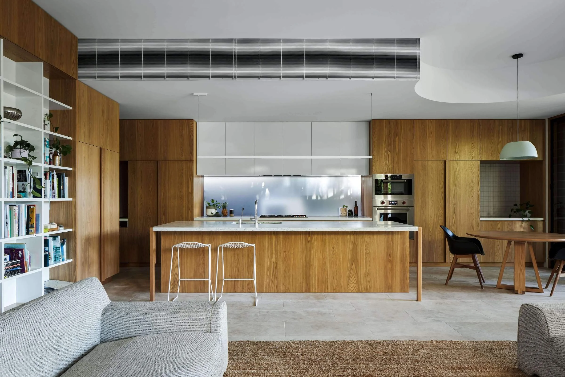 Modern kitchen with wooden cabinets, white countertops, and a gray backsplash, connected to a living space with a gray sofa, a white bookshelf, and a round dining table with black chairs.