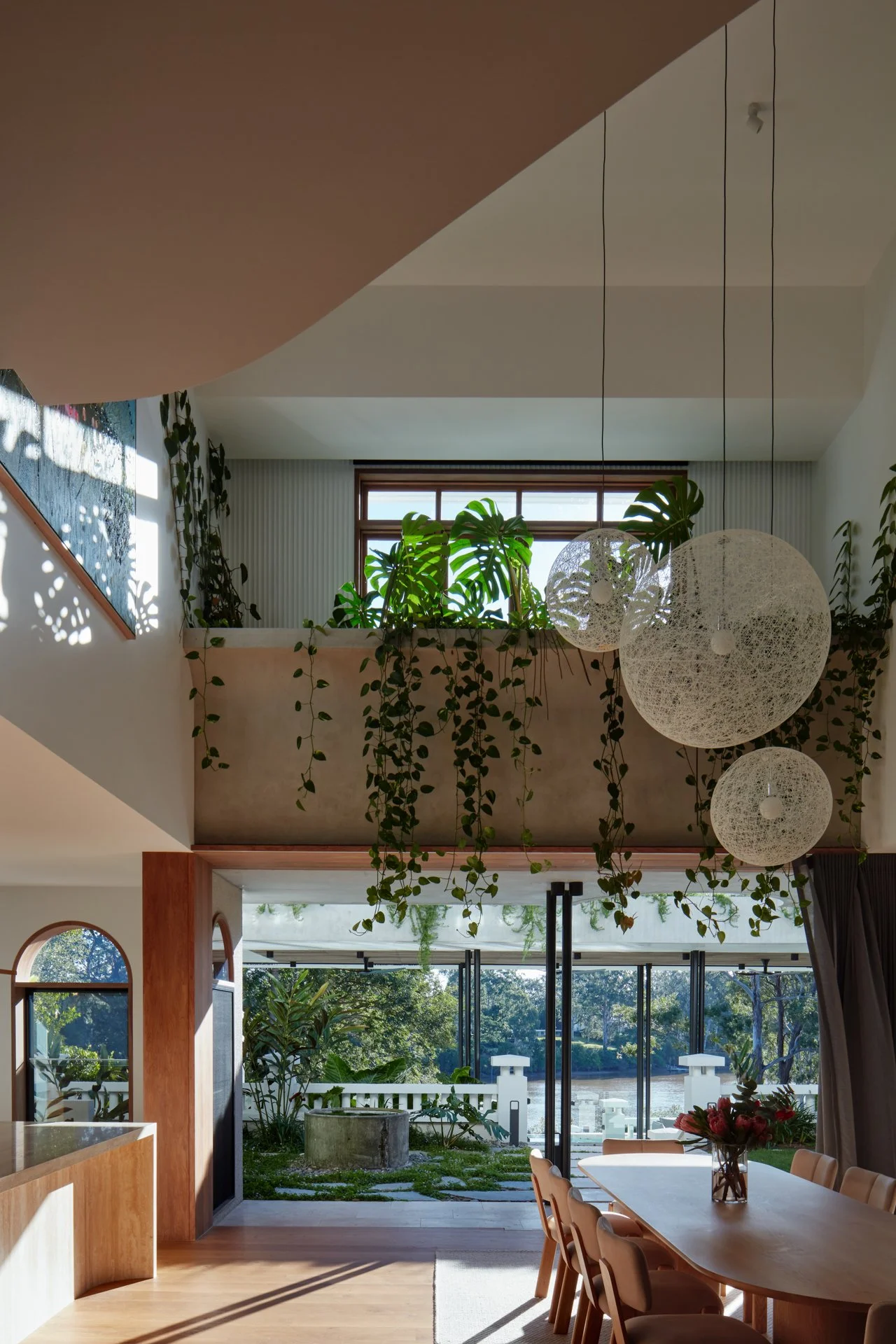 Bright, modern dining room with large windows overlooking a river, wooden dining table with beige chairs, hanging spherical white pendant lights, indoor plants, and natural sunlight.