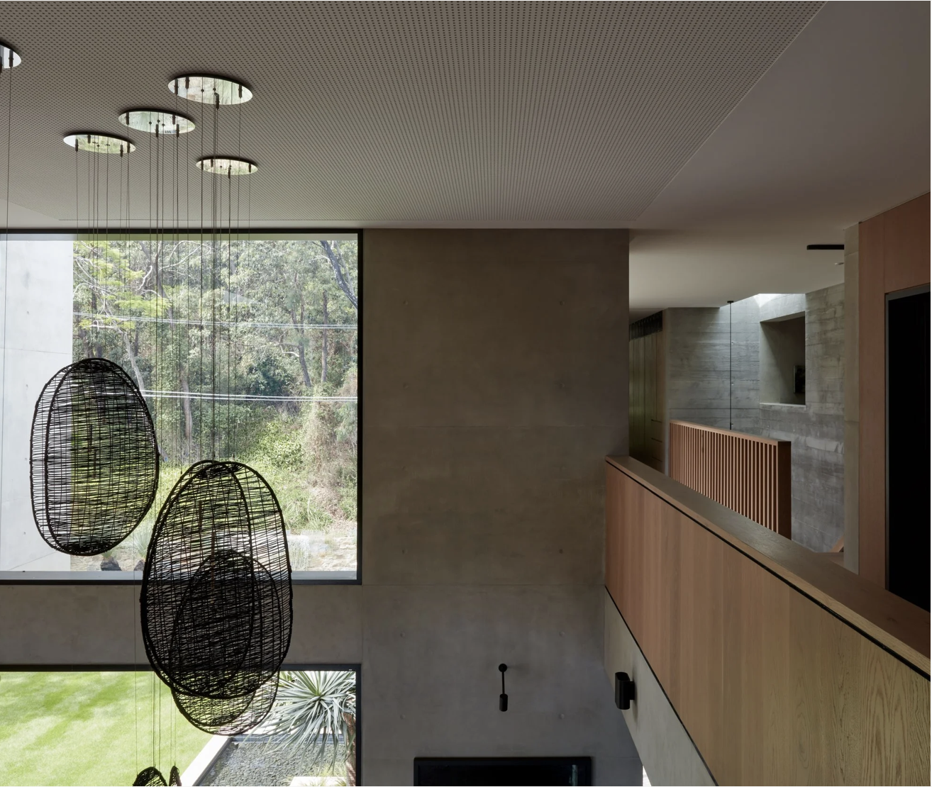 Modern interior with large window view of trees, hanging woven pendant lights, concrete and wood walls, and a railing.