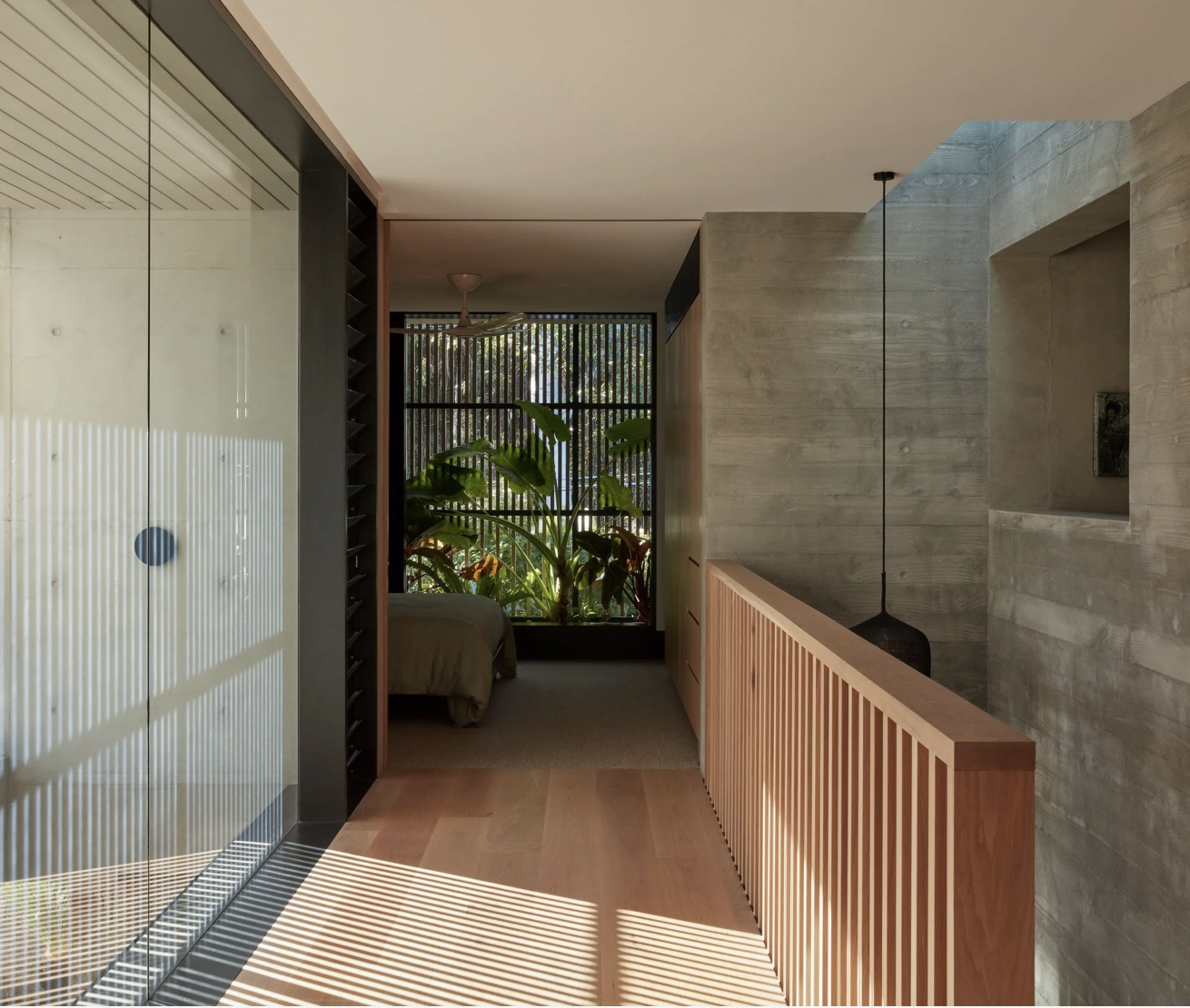 Interior view of a modern bedroom with natural light, wooden accents, and large leafy plants seen through a window, with shadows from blinds on the floor.