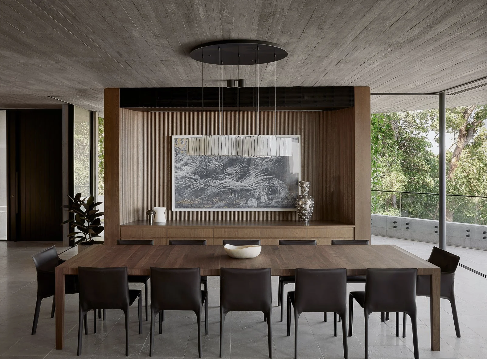 Modern dining room with a large wooden table, black chairs, and a wooden wall with artwork and decorative vases, overlooking a balcony with trees outside.