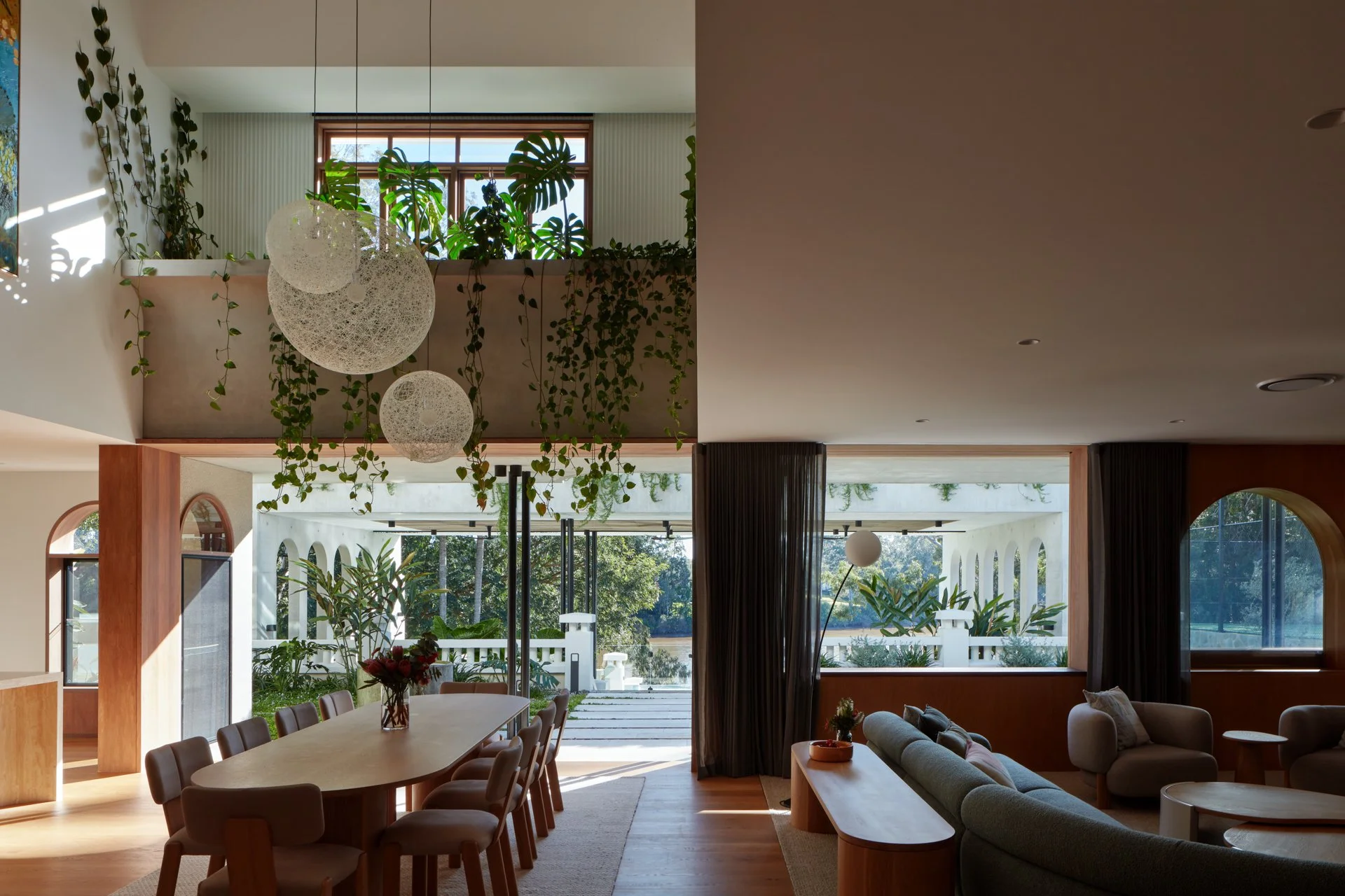Interior of a modern living and dining area with large windows, plants, hanging light fixtures, a dining table with chairs, and a cozy seating area with sofas and a floor lamp.