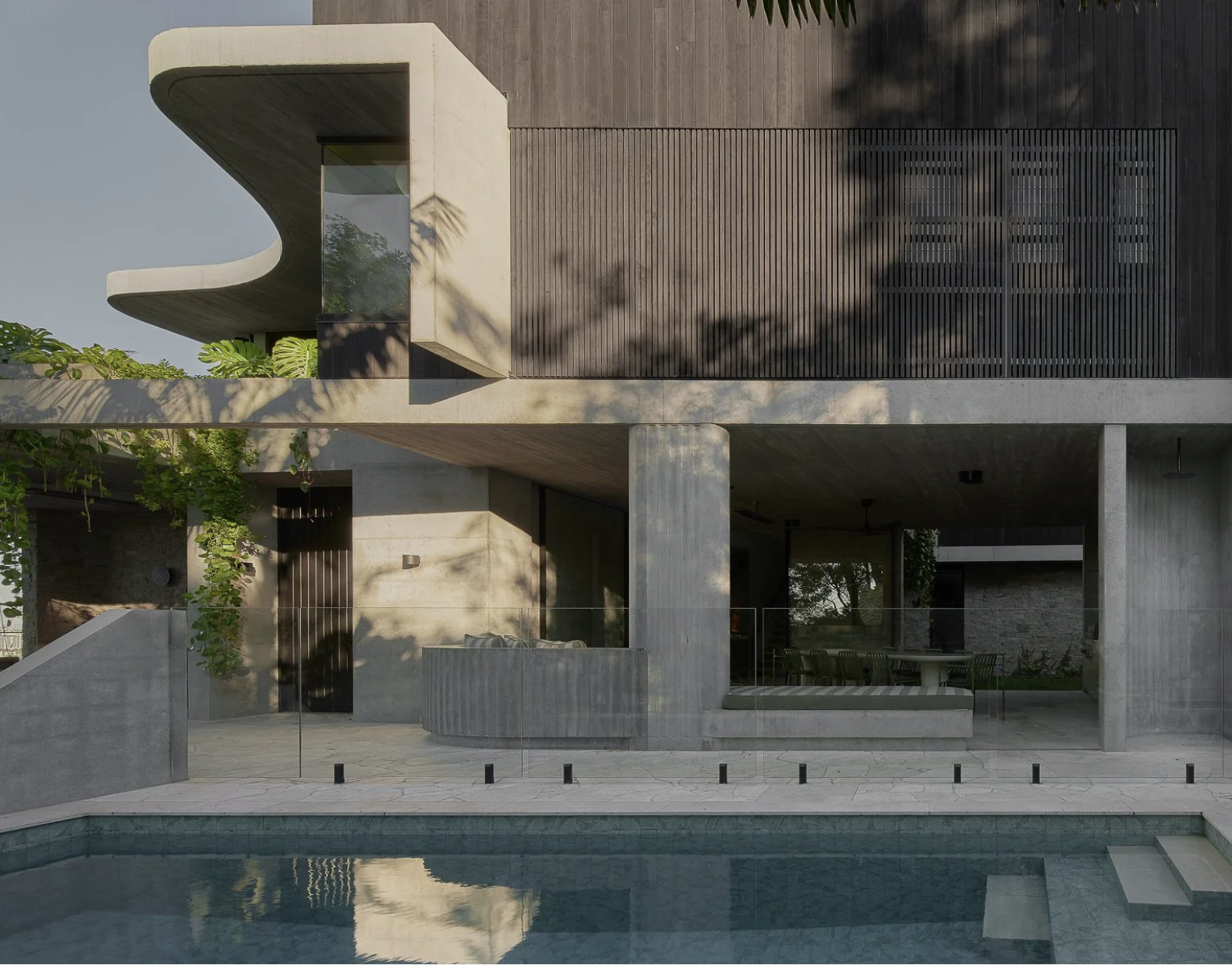 Modern multi-story house with concrete and wooden exterior, pool in the foreground, and shaded outdoor seating area.