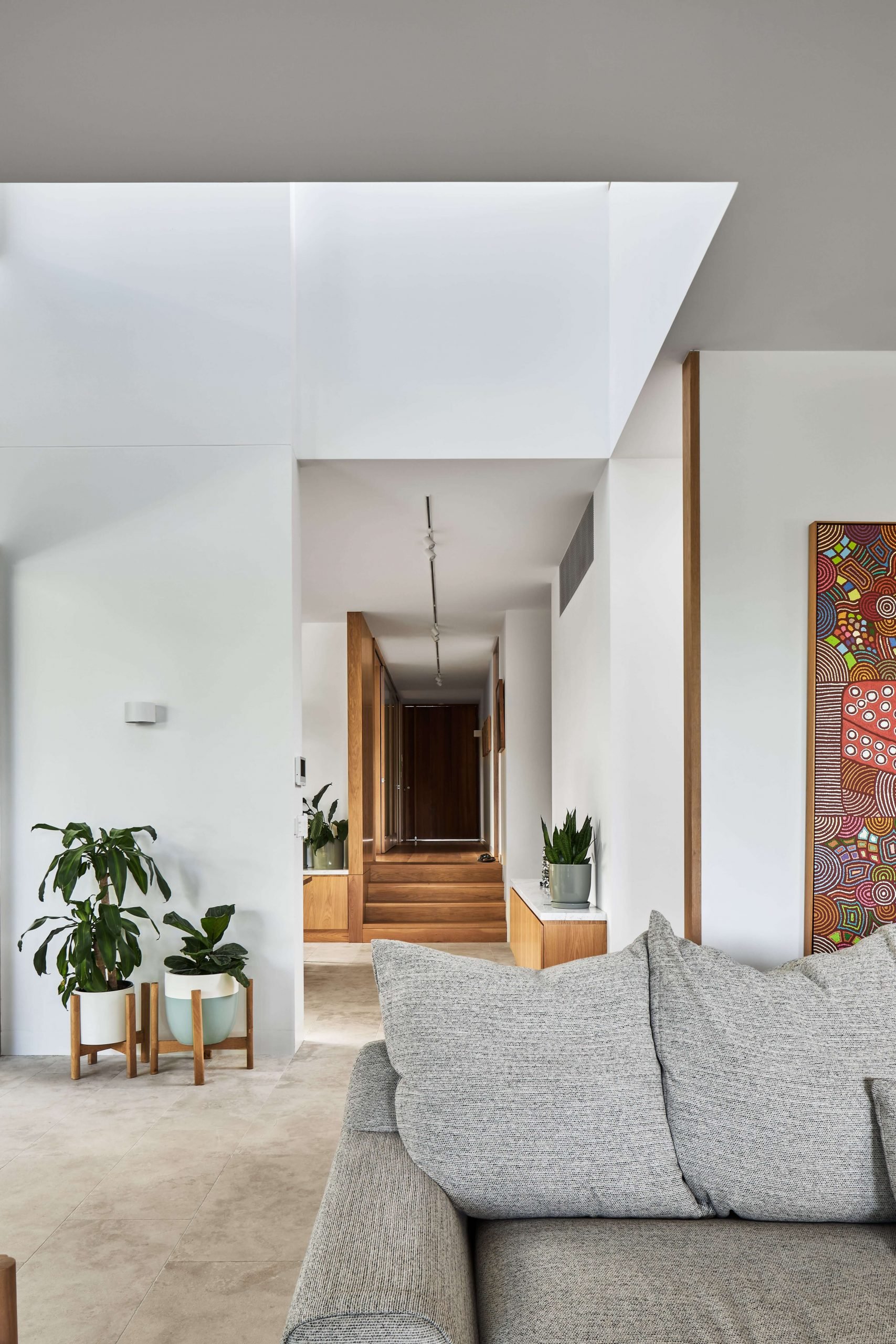Interior of a modern living room with a gray sofa, houseplants in white pots on wooden stands, a colorful abstract painting, and a hallway with wooden stairs leading upstairs.